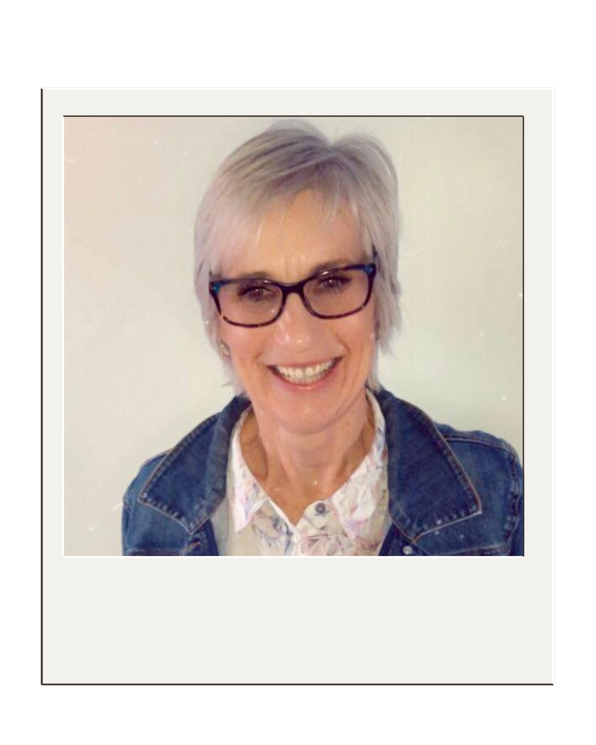 A woman with short gray hair and glasses smiling, wearing a denim jacket and a floral shirt, standing against a plain light-colored wall.