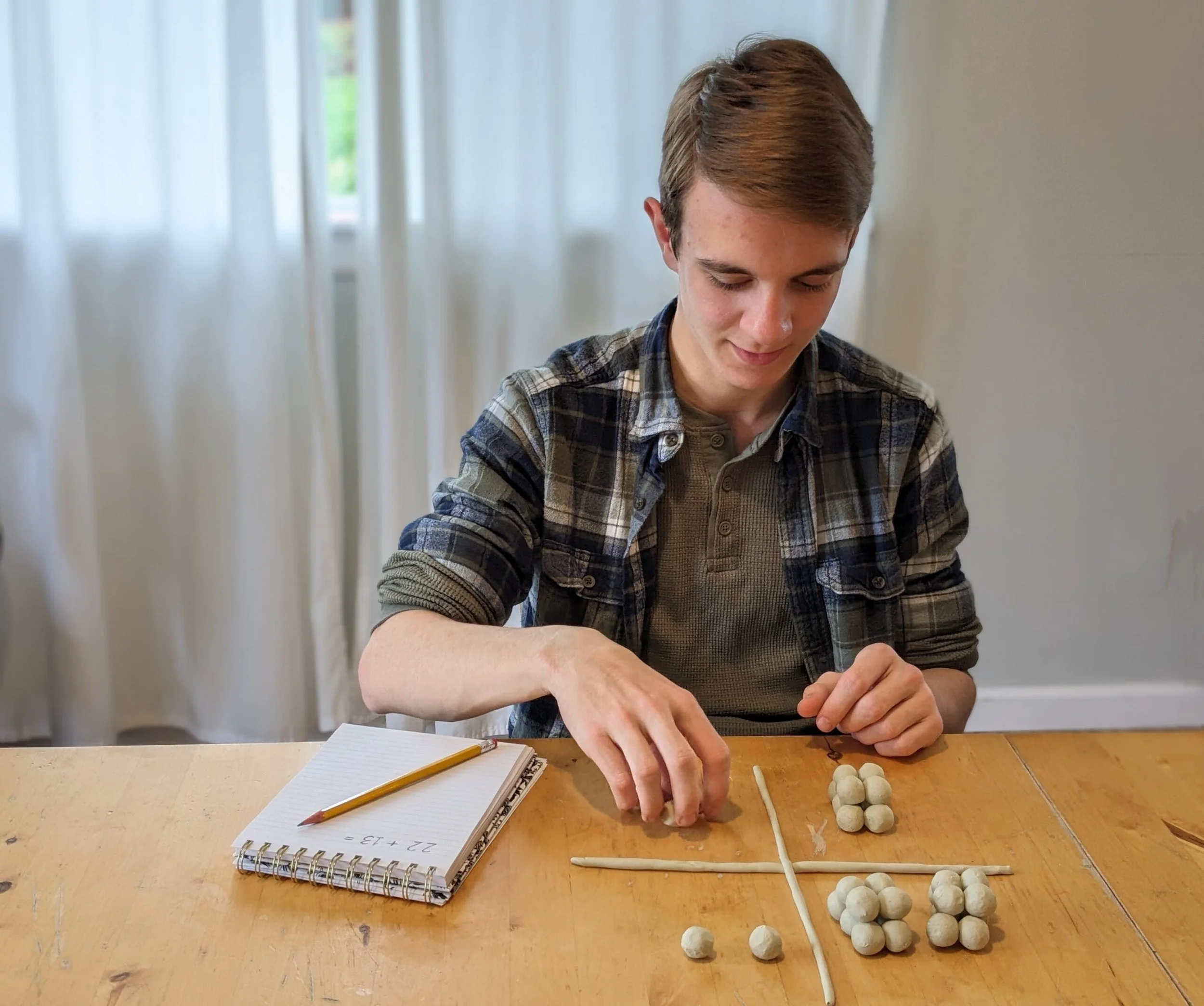 A young man with brown hair wearing a plaid shirt working on a project involving small spherical objects on a wooden table, with a notebook, pencil, and notepad nearby, in a room with white curtains and natural light.