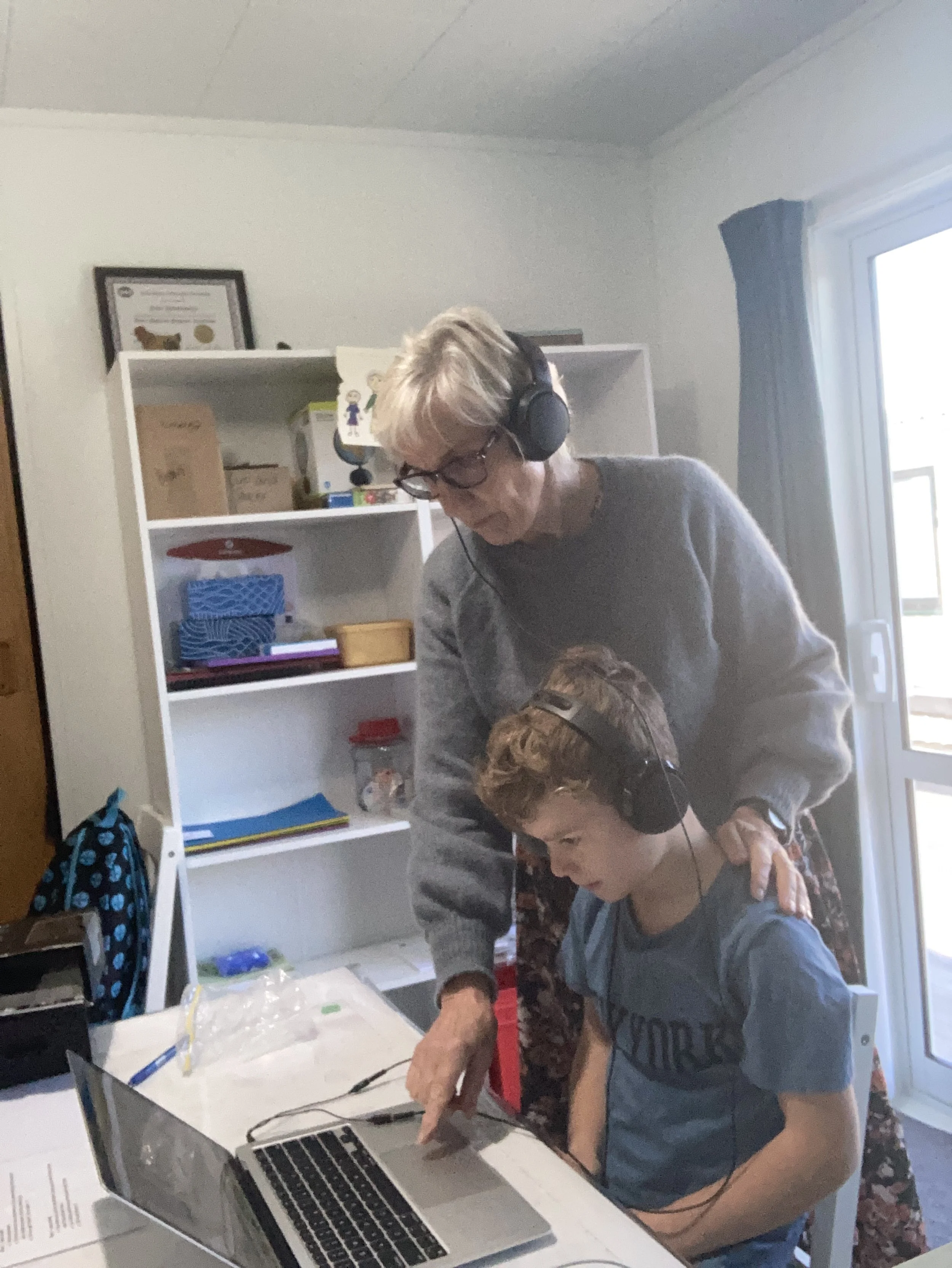An older woman and a young boy are working together on a project at a table. The woman wears headphones and glasses and is leaning over, while the boy is sitting and looking at the project. There is a white table with a laptop, papers, and some supplies in the room with a bookshelf and a window with curtains.