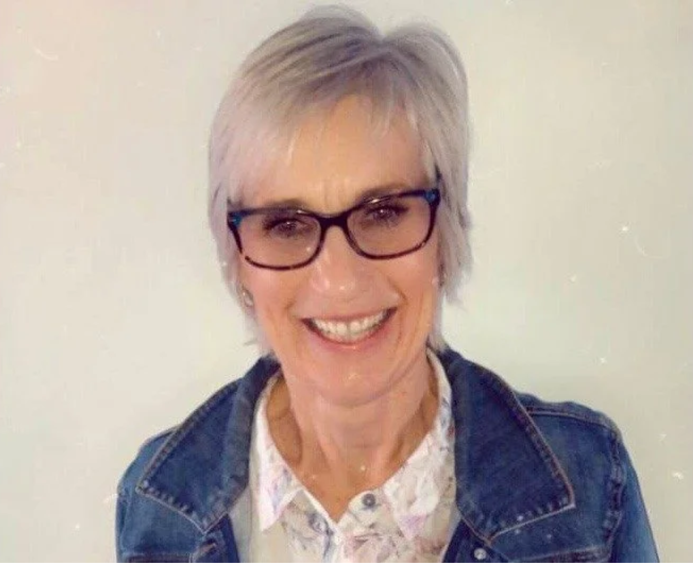 A smiling woman with short gray hair and glasses, wearing a dark jacket over a white shirt, standing against a plain light-colored background.