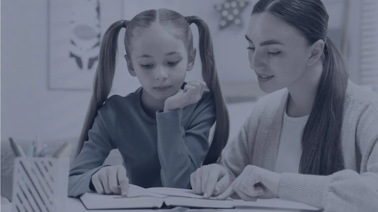 A woman helps a young girl with her homework at a table.