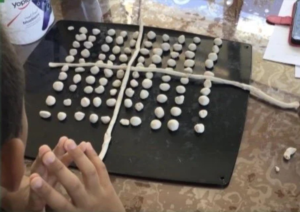 Person making a shape with small dough balls and two long strands of dough on a black surface, with a measuring tape in the background.