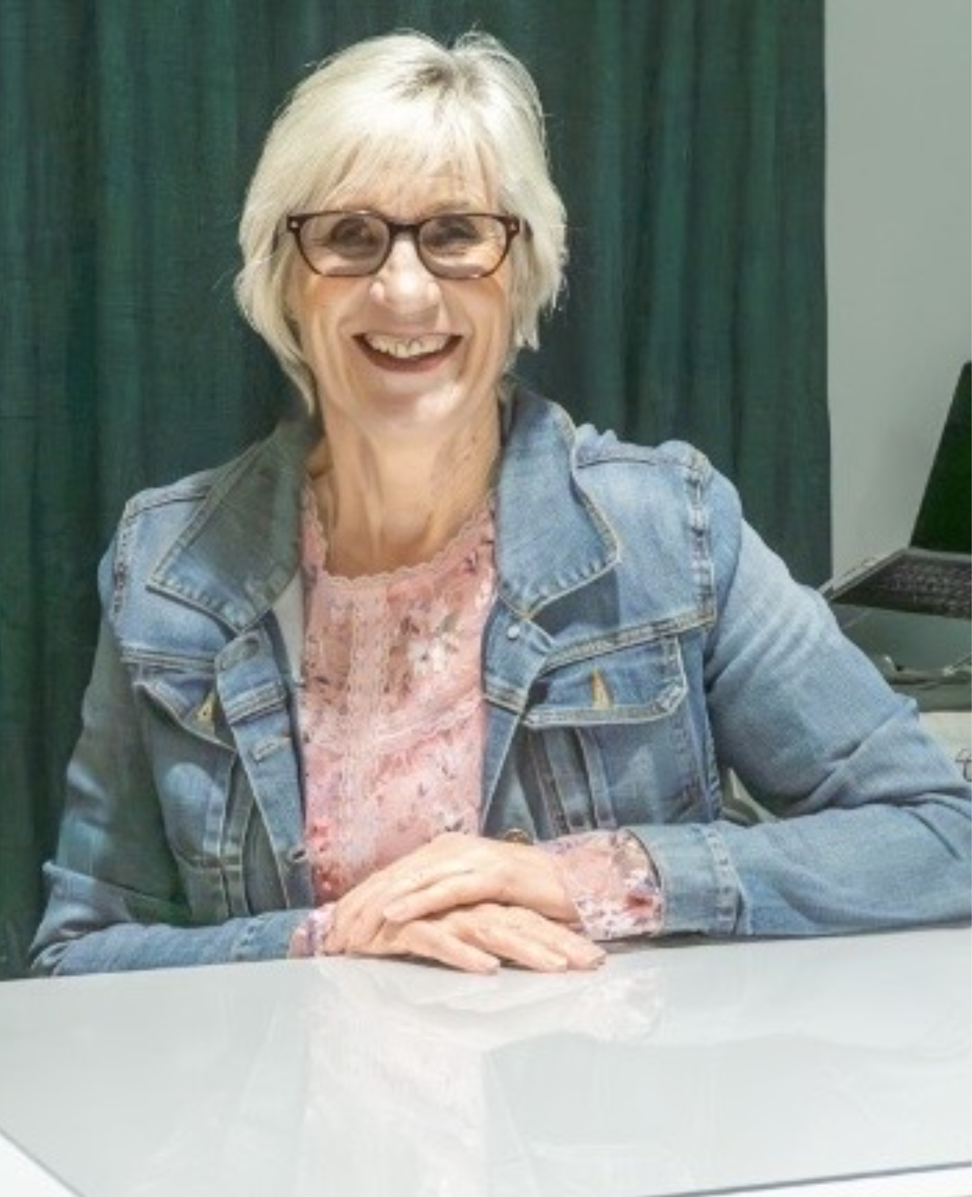 A woman with short blonde hair, glasses, and a denim jacket smiling at the camera, sitting at a table with a laptop in front of her, against a dark curtain background.