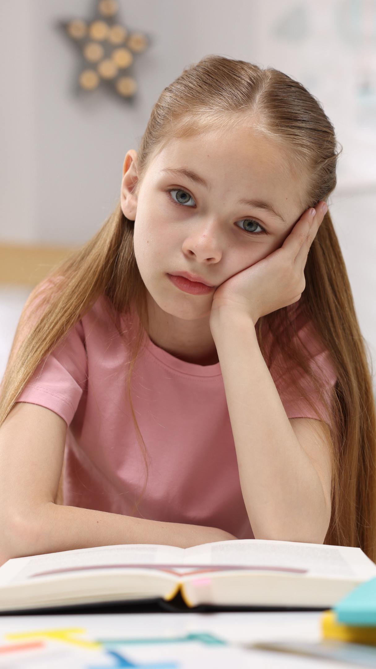 A young girl with long, light brown hair and blue eyes rests her face on her hand while seated at a desk with an open book and colorful sticky notes.