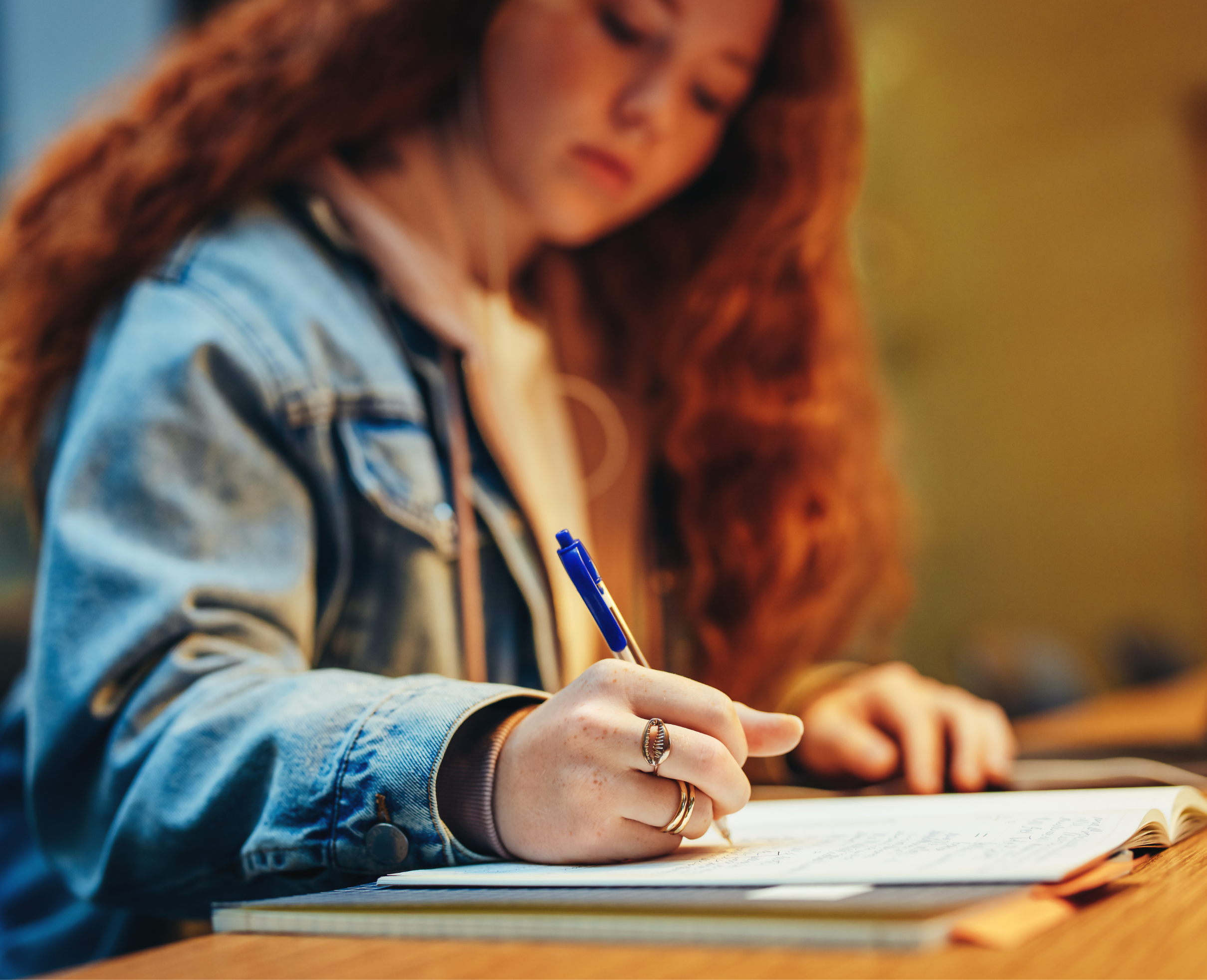 A young woman with curly red hair wearing a denim jacket, writing in a notebook with a blue pen.