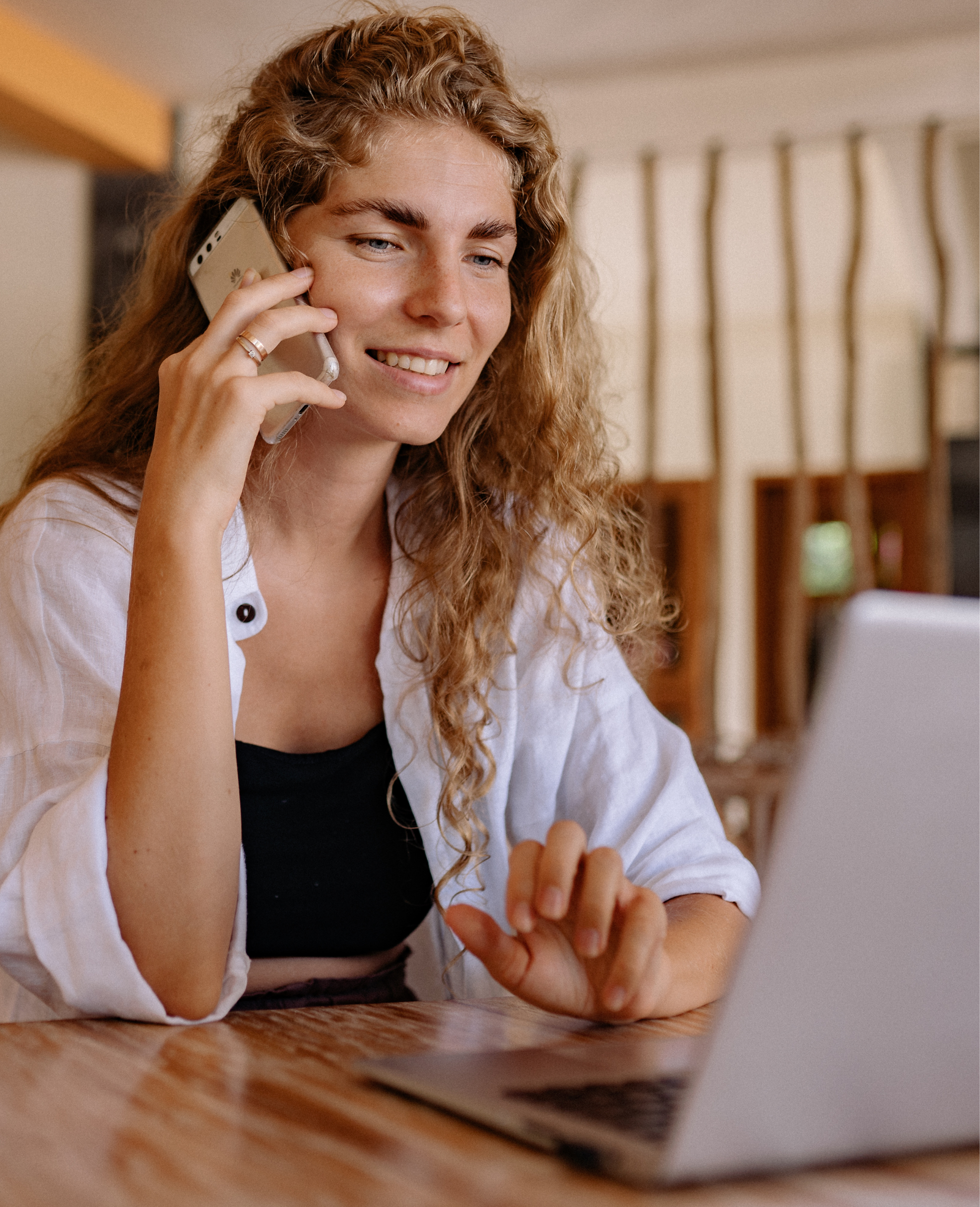 A woman with curly blonde hair is talking on a landline phone while looking at her laptop on a wooden table.