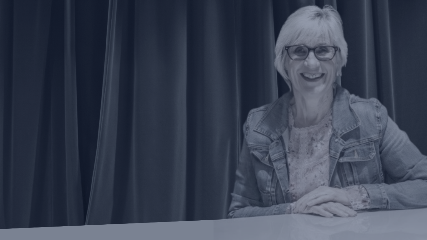 A smiling woman with blonde hair, glasses, and a denim jacket sitting at a table with dark curtain background.