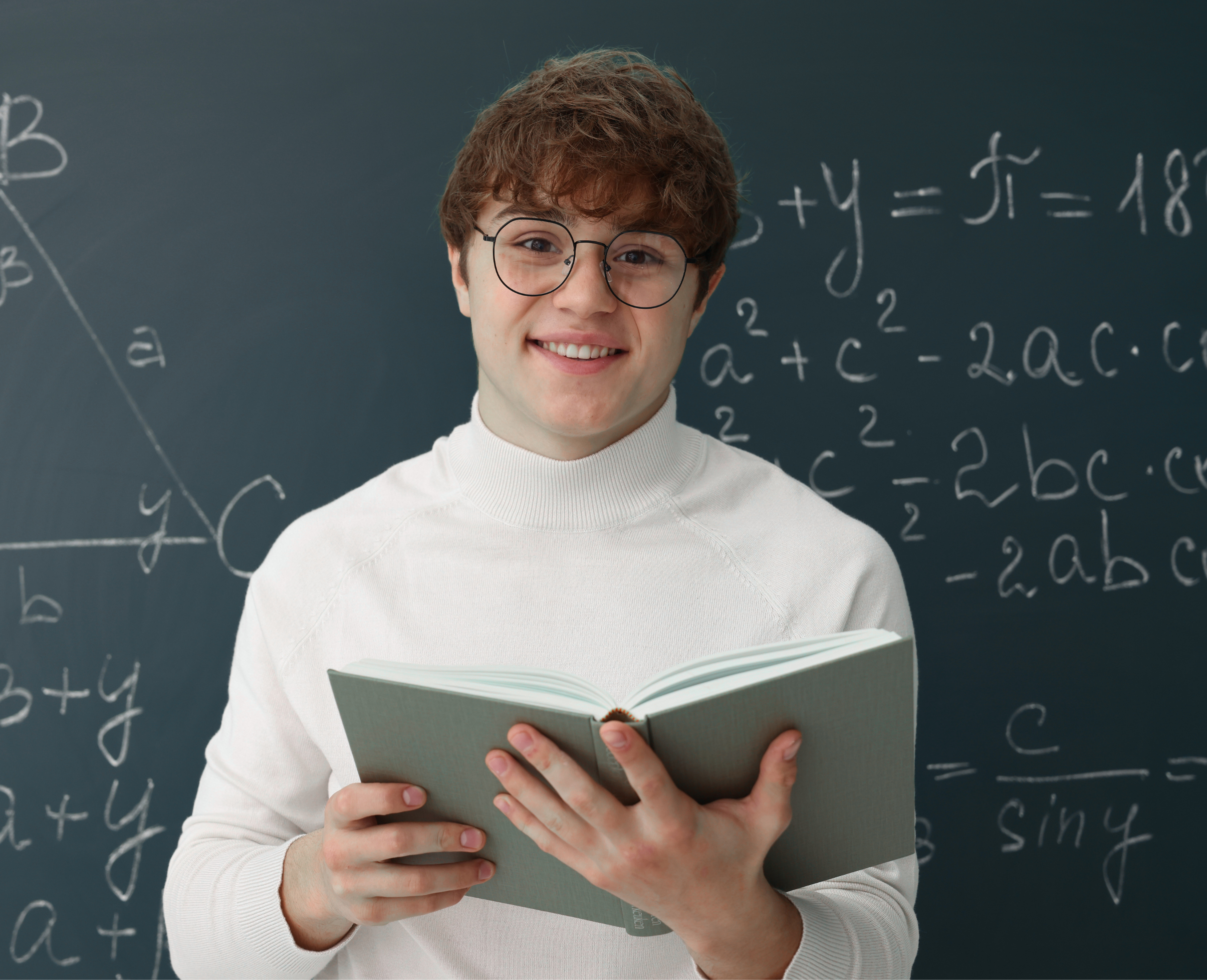 A young man with curly brown hair, glasses, and a white sweater reading a book in front of a blackboard filled with mathematical equations.