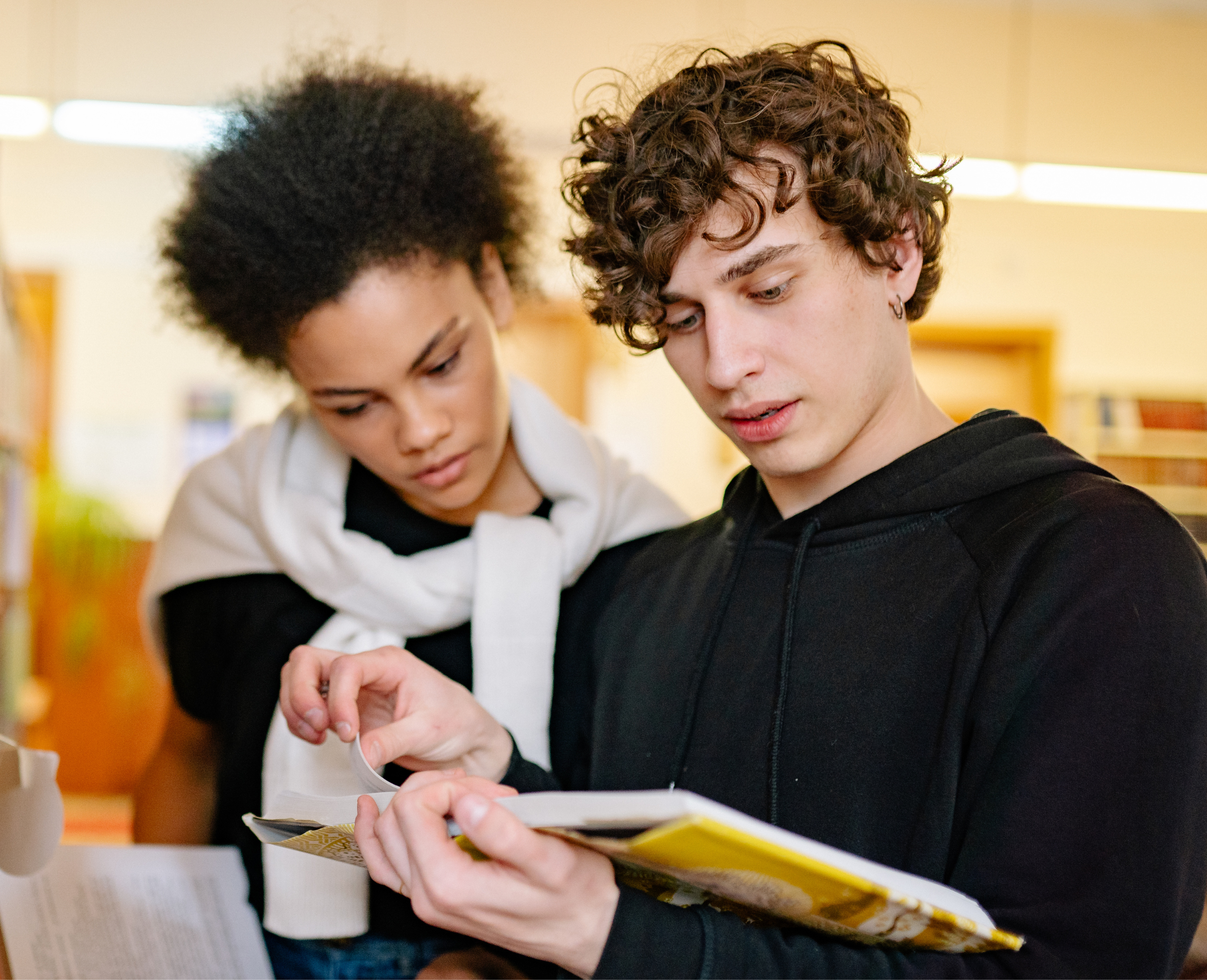 Two young people, a man with curly brown hair and a woman with short, natural black hair, are looking at an electronic device together, possibly a phone or tablet, in a well-lit indoor environment.