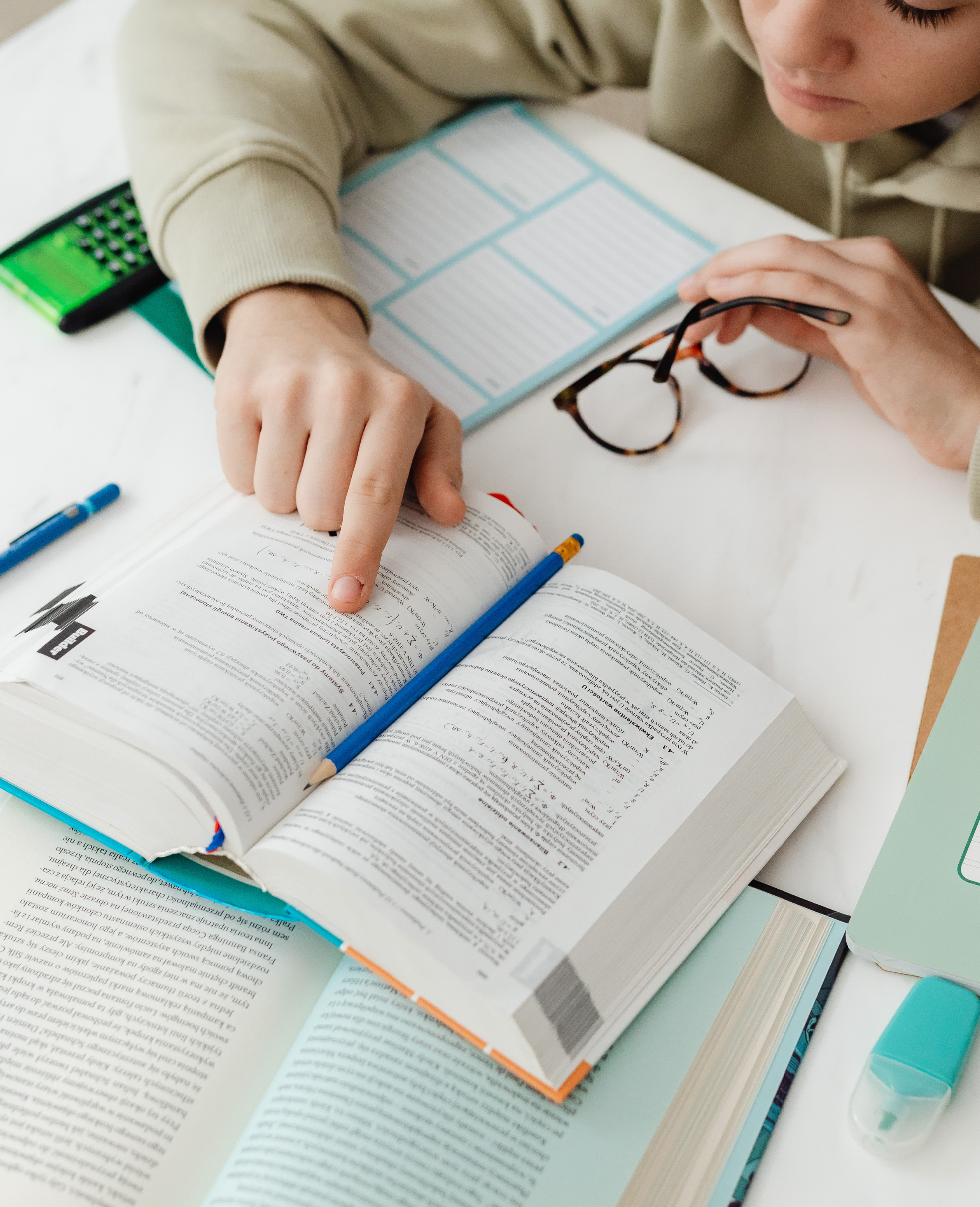 Open textbook with highlighted text, a blue pen, glasses, a multicolored highlighter, a calculator, and a bottle of hand sanitizer on a white table.