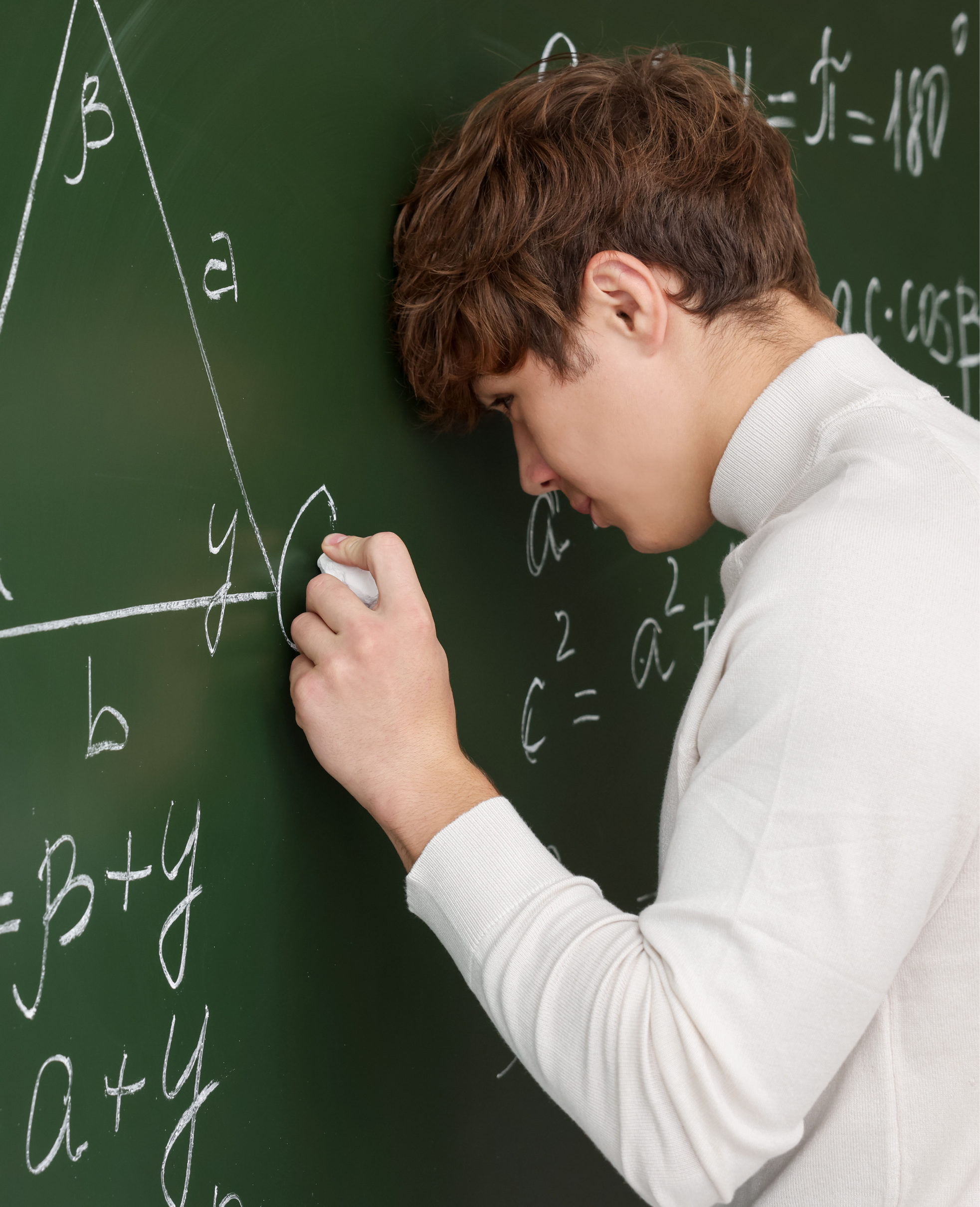 A student writing on a green chalkboard filled with math and science equations and diagrams, wearing a white long-sleeve shirt.
