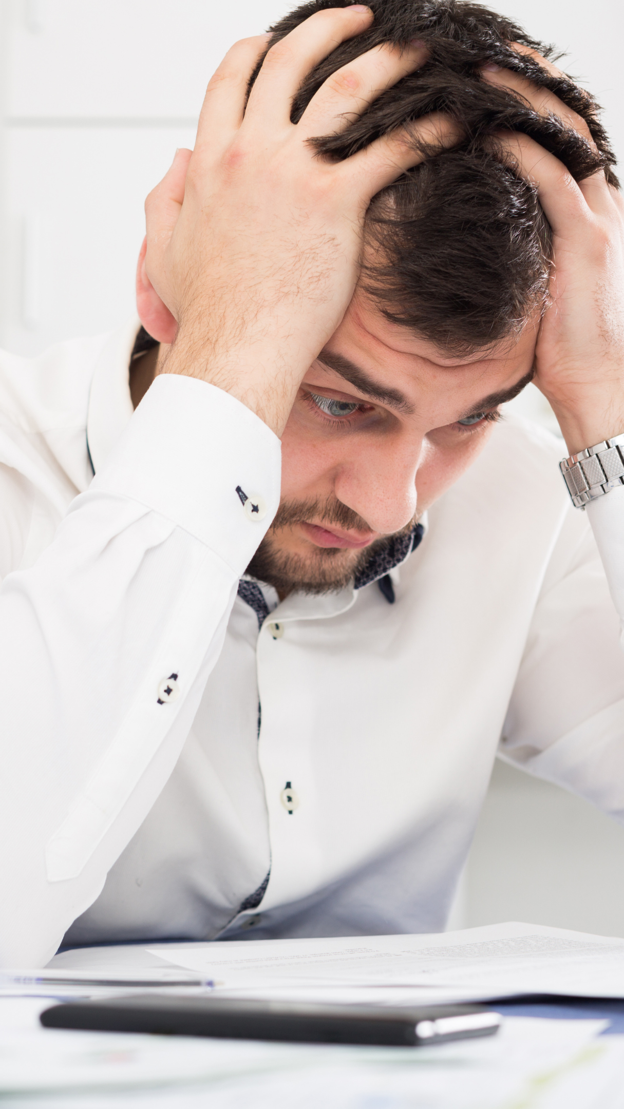 A man with dark hair and a beard, wearing a white shirt, sitting at a desk with a pen and paper, holding his head with both hands, looking down with a stressed expression.