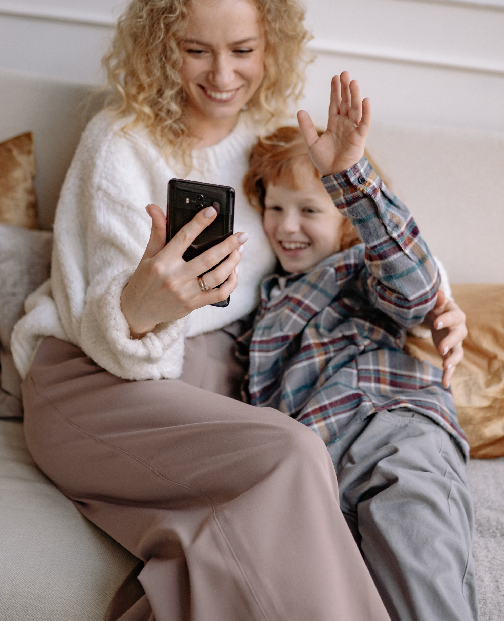 A woman with curly blonde hair and a boy with red hair sitting together on a beige sofa. The woman is holding a smartphone, smiling, and the boy is waving at the camera. The woman is wearing a white sweater and light brown pants, the boy a plaid shirt and gray pants.