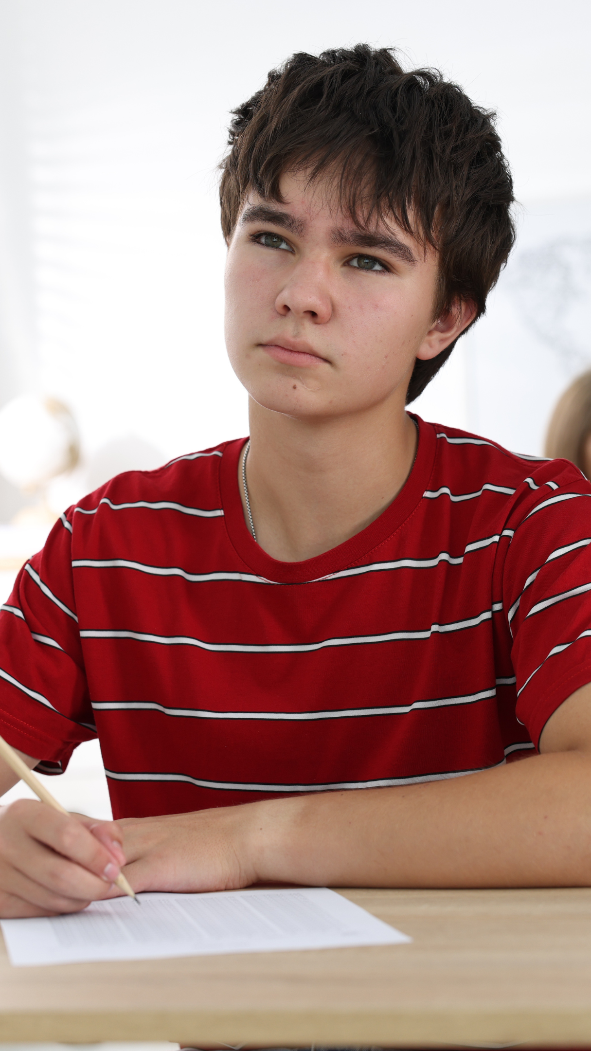 A teenage boy with dark hair and light skin sitting at a table, holding a pencil and writing on a sheet of paper, wearing a red and white striped t-shirt, in a bright room.