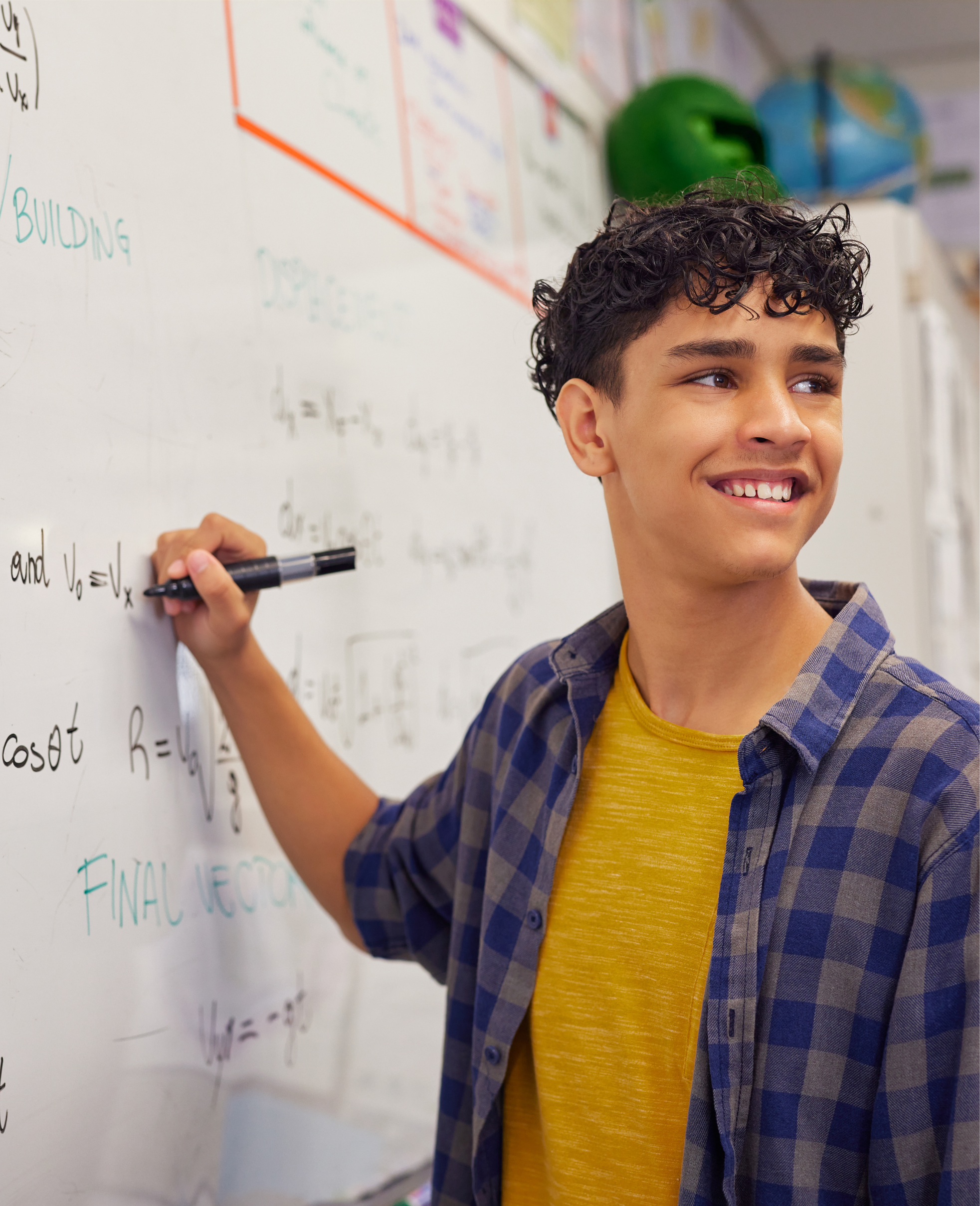 A young person with curly hair, wearing a yellow T-shirt and a blue plaid shirt, writing on a whiteboard with a black marker, smiling, in front of a wall with colorful educational posters and a green and blue globe.