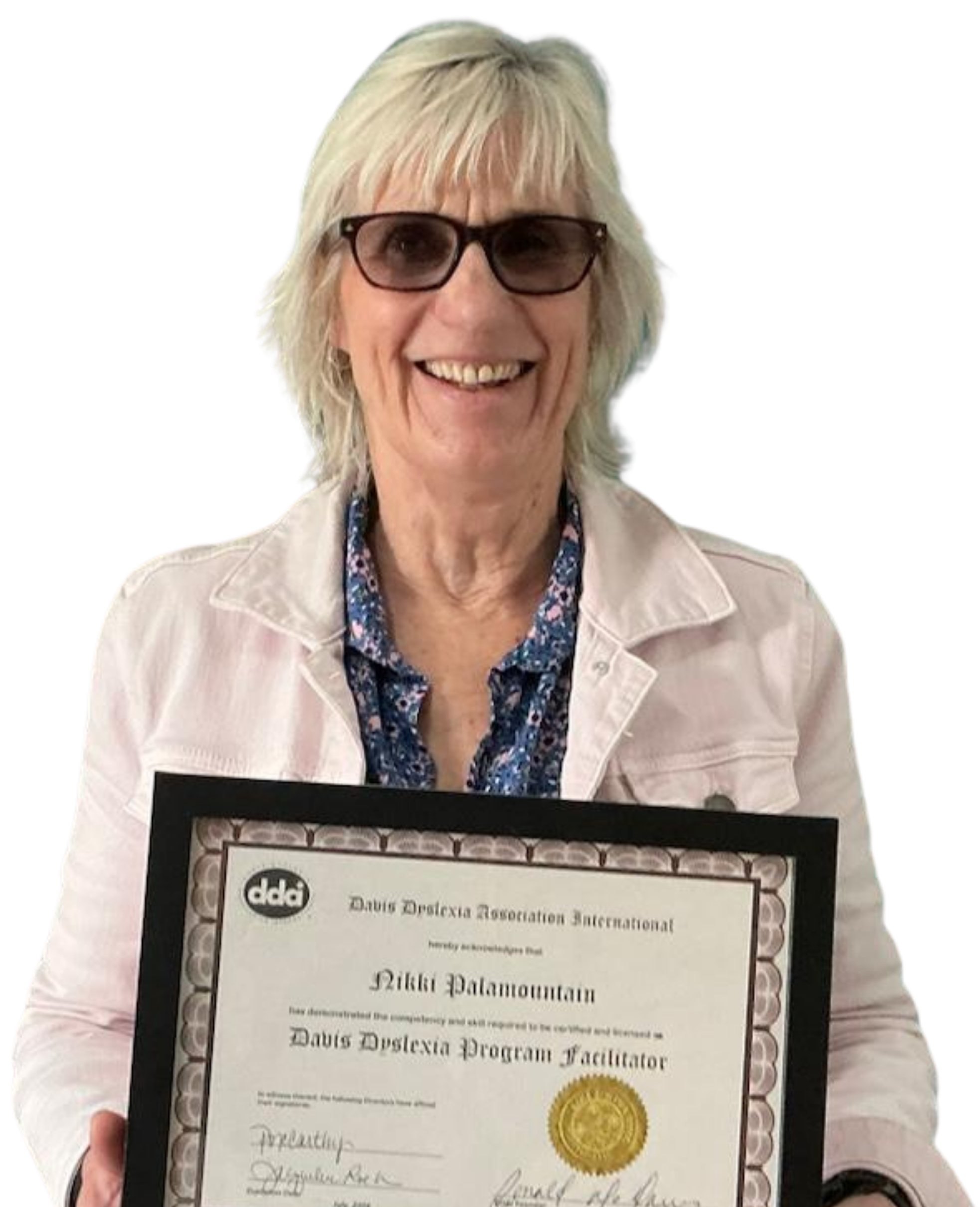 A woman with blonde hair and glasses holding a diploma certificate. She is smiling and wearing a light-colored jacket over a patterned blouse.