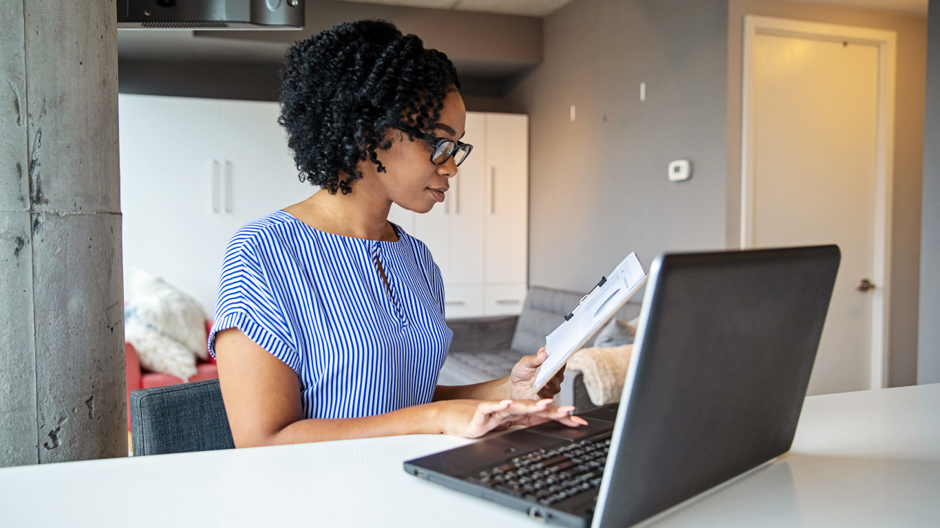 A woman with glasses and curly hair sitting at a table, looking at a paper while working on a laptop in a modern home kitchen.