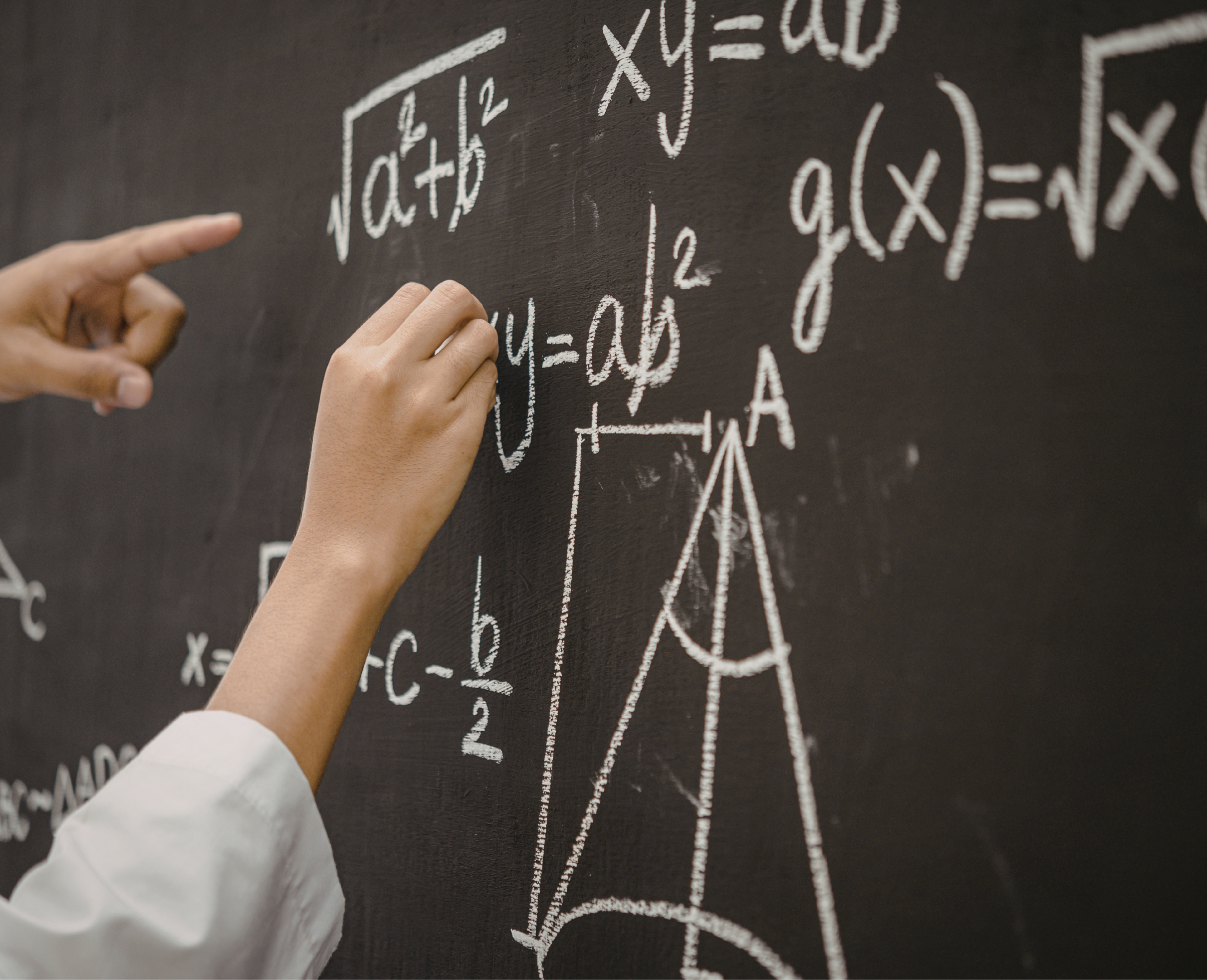 A person's hand writing mathematical formulas and equations on a black chalkboard with white chalk, including symbols and variables.