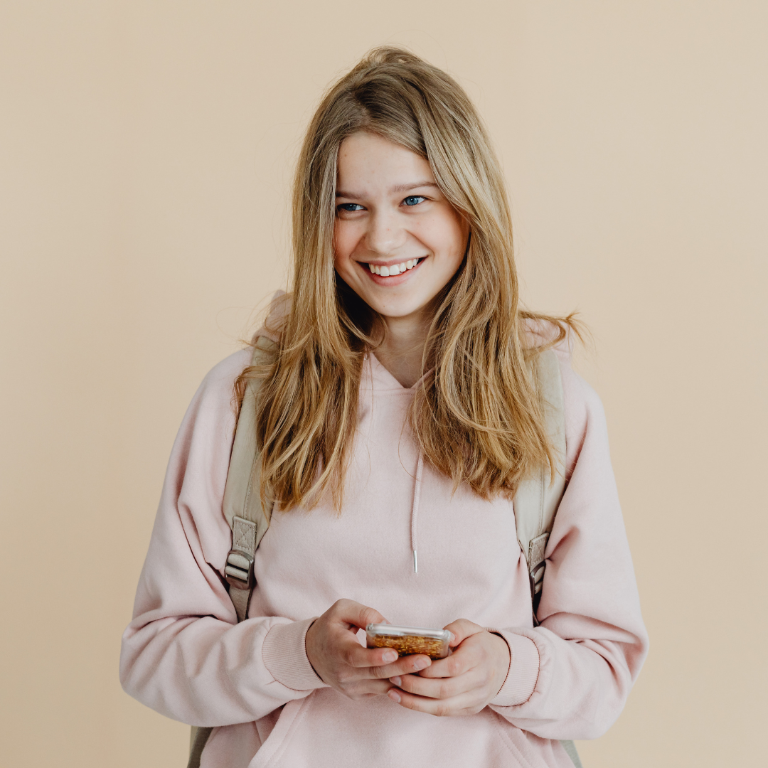 A young woman with long blond hair, smiling, looking to her right, wearing a light pink hoodie and carrying a backpack, holding a smartphone, standing against a plain beige background.