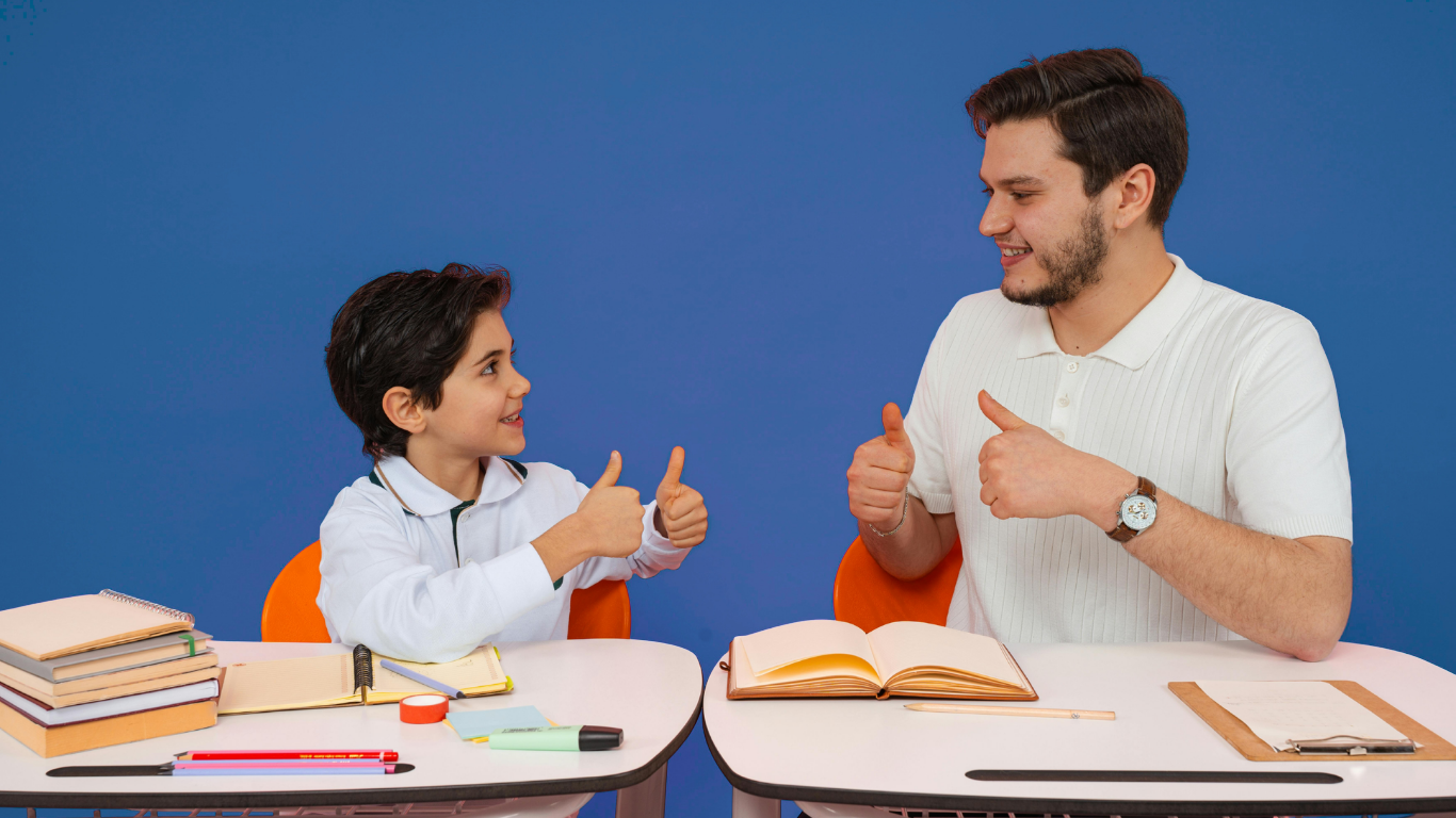 A young boy and an adult man sitting at tables facing each other, smiling, with their thumbs up in a friendly gesture against a blue background. The tables have books, notebooks, pens, and other school supplies.