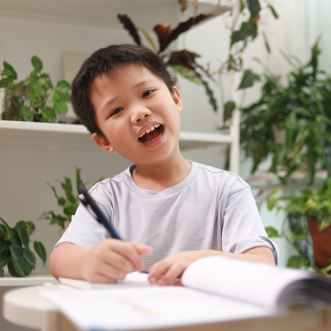 A young boy smiling and writing in a notebook at a desk surrounded by green plants.