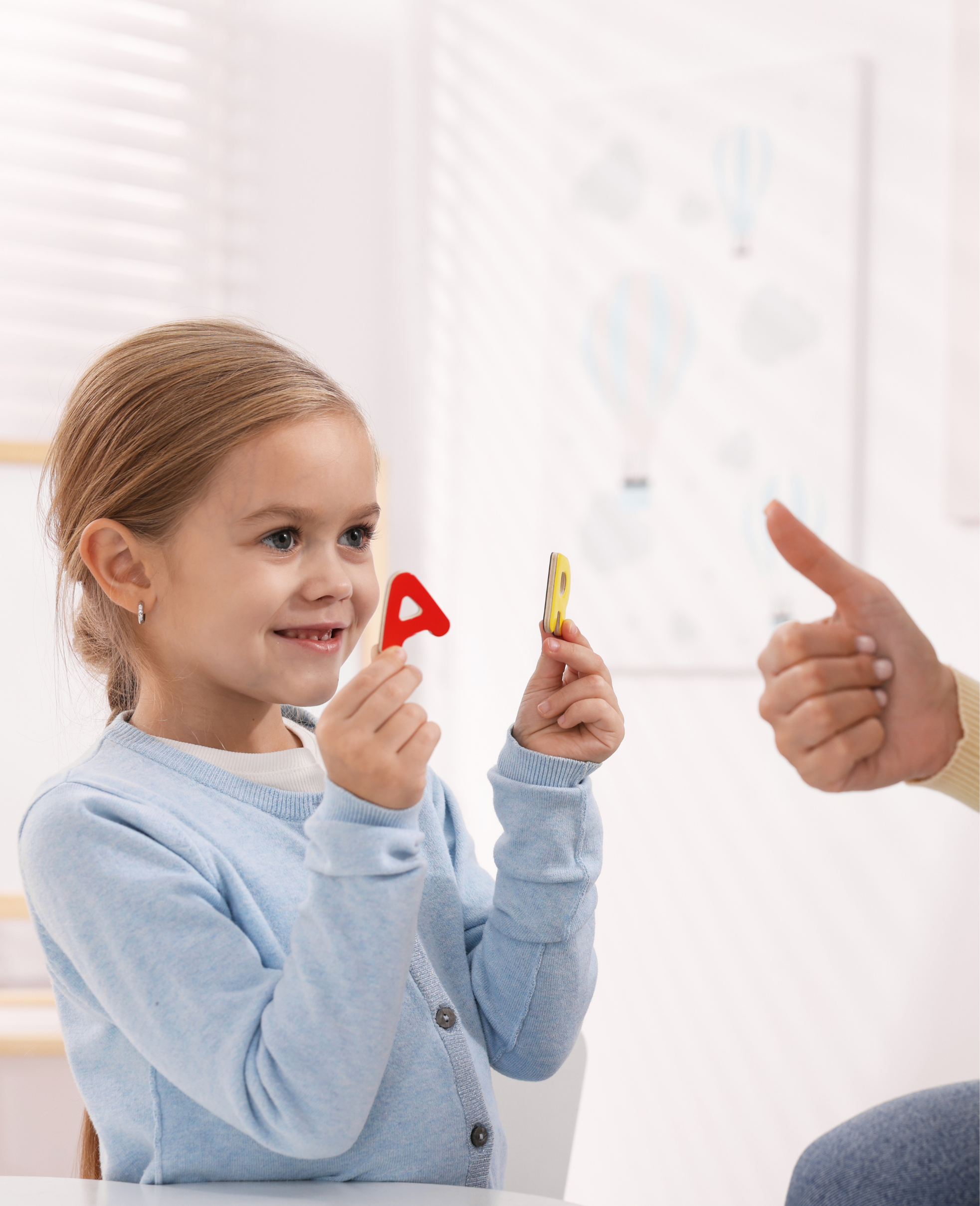A young girl with blonde hair and blue eyes holding red and yellow alphabet letters, engaged in a discussion with an adult pointing at the girl, with a blurred educational poster in the background.