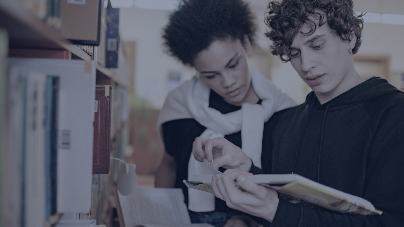 Two young adults with curly hair reading a book together at a library.