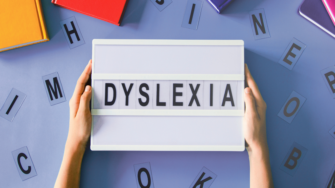 Hands holding a letter board with the word "DYSLEXIA" on it, surrounded by scattered alphabet cards on a blue surface.