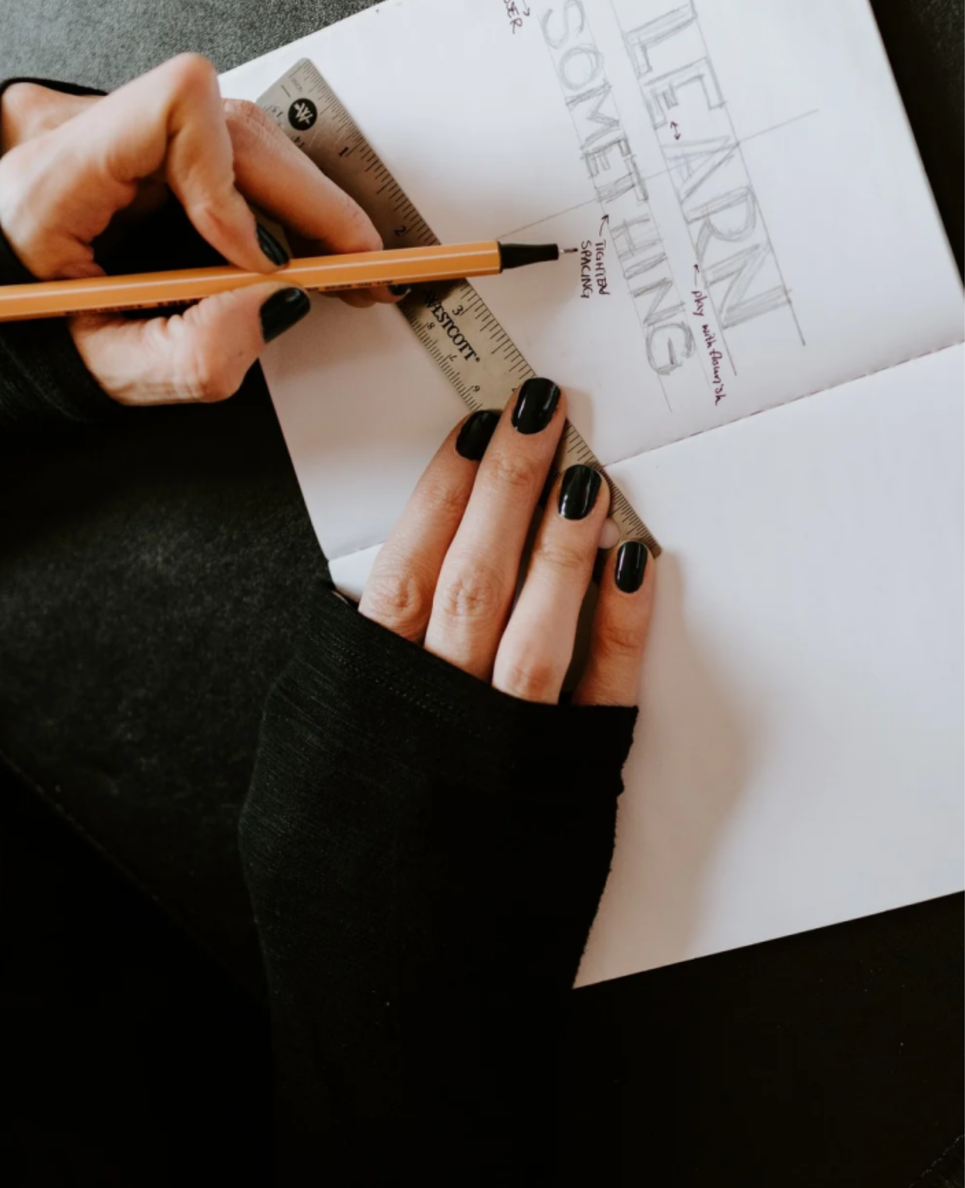 Close-up of a person's hands with black nail polish holding a ruler and a pen, drawing on a sheet of paper with sketches and notes, including the word 'ARCHITECTURE' and related annotations.