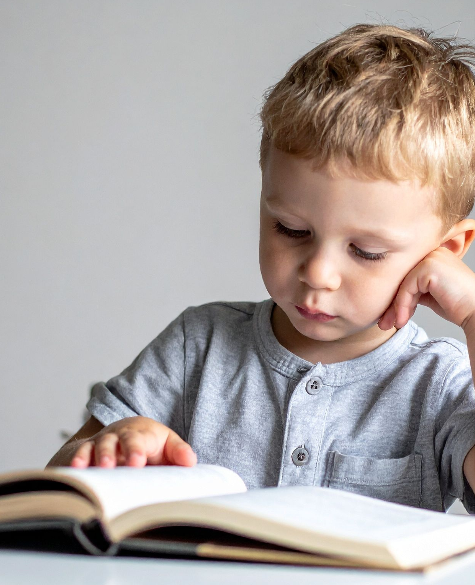 A young boy with curly red hair and wearing a gray shirt, looking down and touching his face with his right hand, with an open book in front of him.