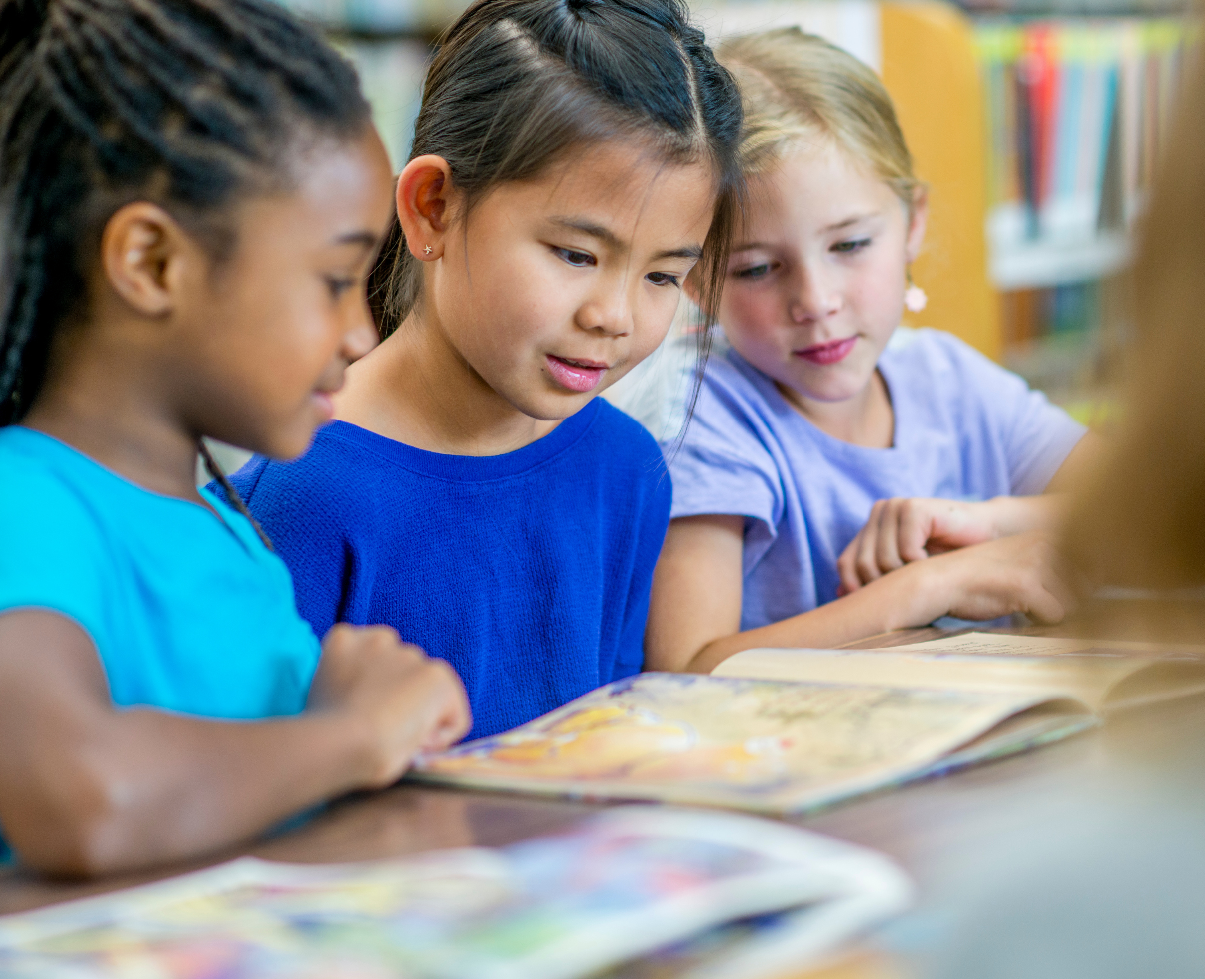 Four young girls with different hair types and colors are closely looking at and engaging with a book or printed material on a table in a classroom or library setting.