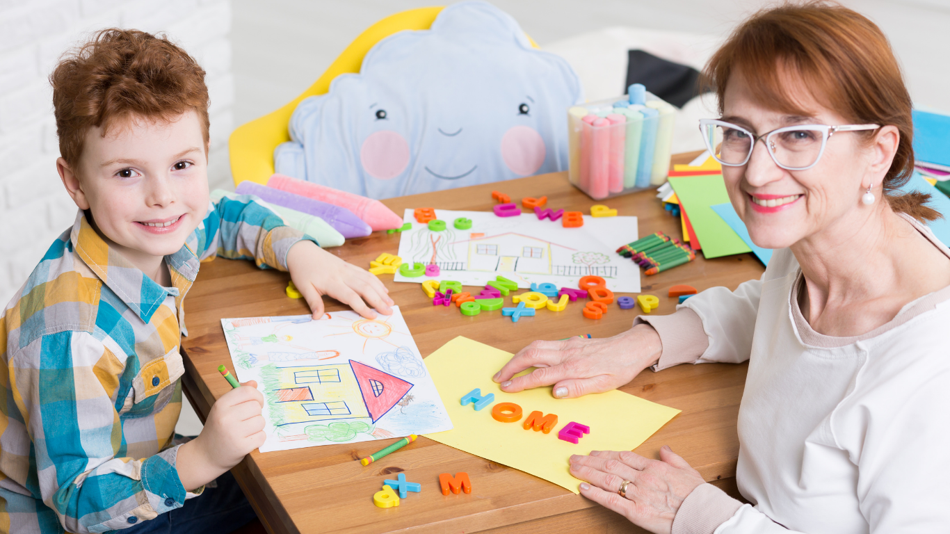 A woman and a young boy sitting at a table engaged in arts and crafts. The table has colorful foam letters, drawings, and craft supplies. There is a cloud-shaped pillow with a smiling face in the background.