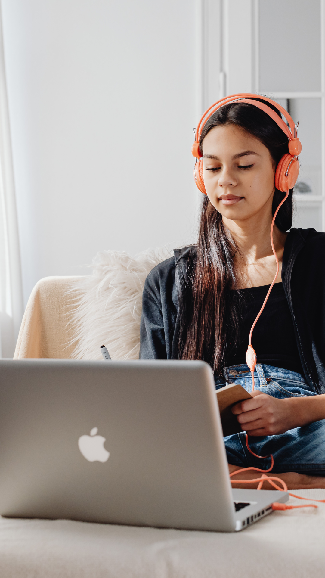 Young woman with long dark hair wearing pink headphones, sitting on a beige couch with a laptop in front of her, holding a small notebook and pen, in a bright room with white walls.