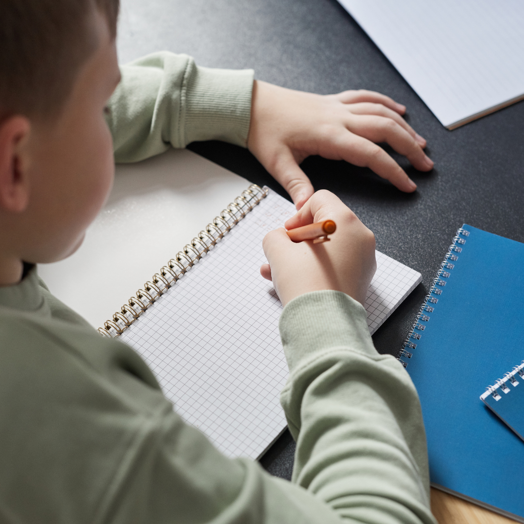 A child writing in a graph paper notebook at a desk with other notebooks and a spiral-bound notepad around.