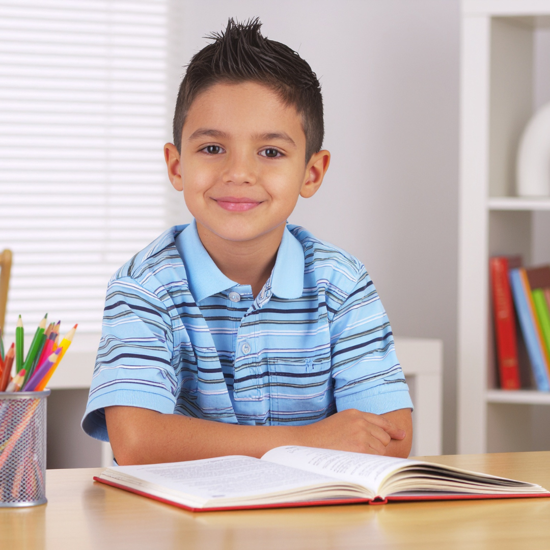 A young boy with short, spiked dark hair smiling and sitting at a desk with an open book in front of him. There is a container of colored pencils on the desk and books on shelves behind him.