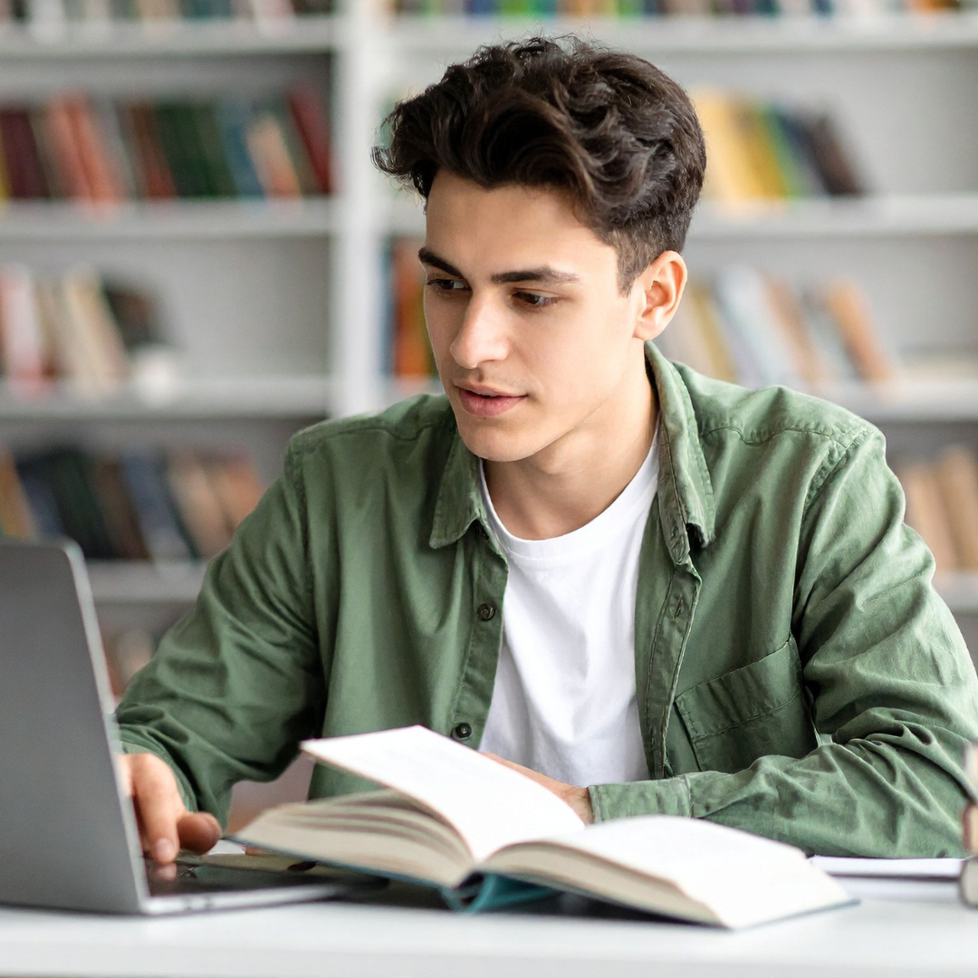 Young man studying with an open book and laptop in a library.