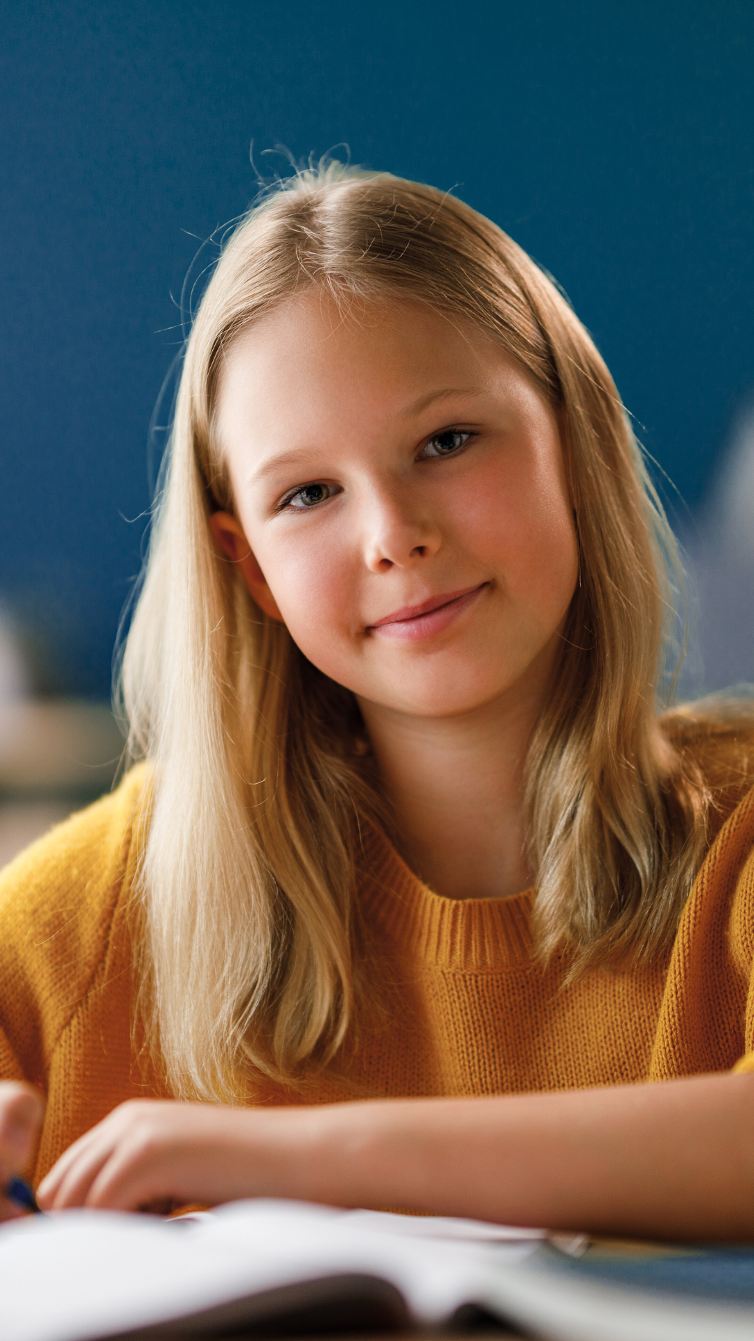 A smiling young girl with long blonde hair in a yellow sweater, sitting at a table with an open book in front of her.