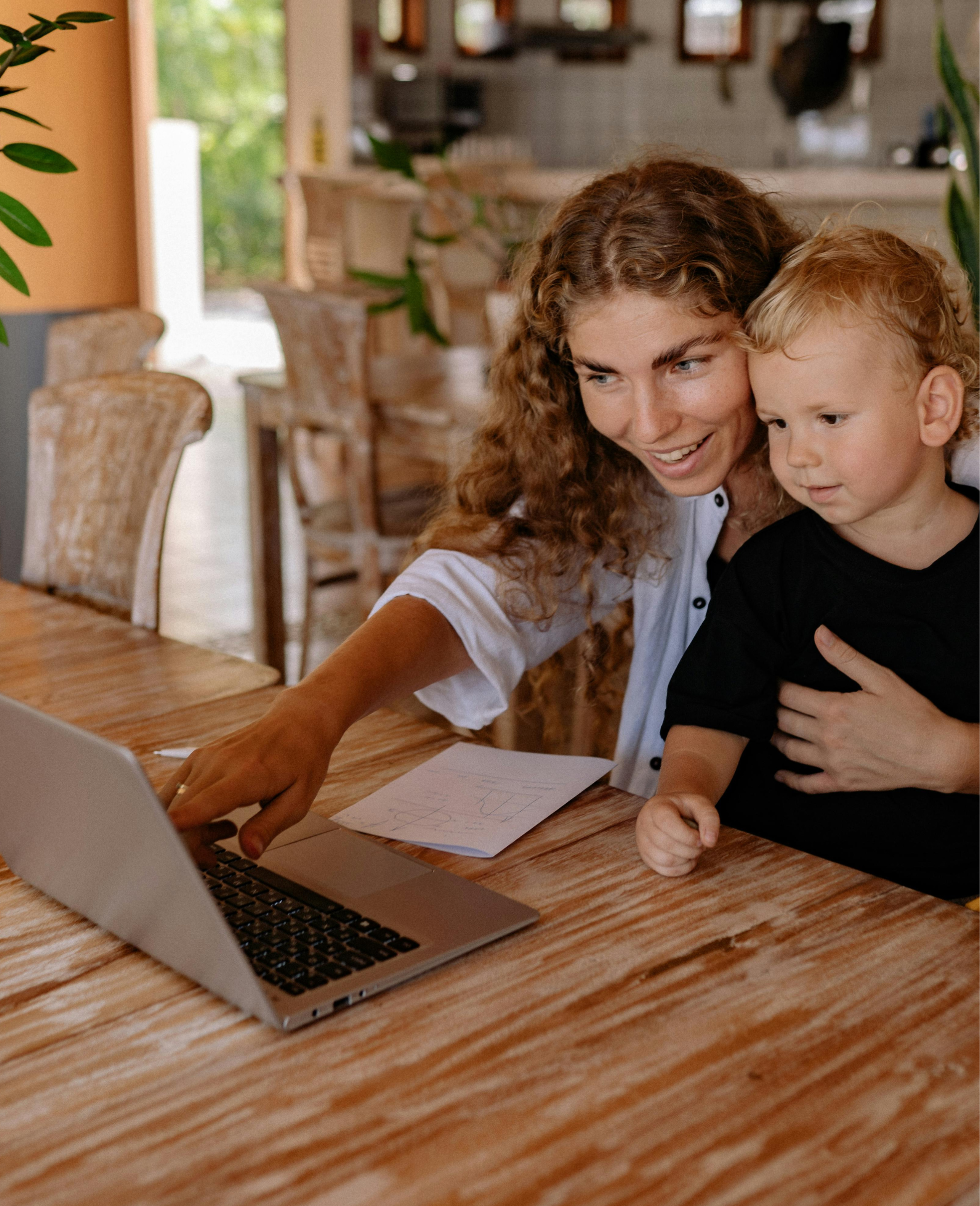 A woman with curly hair and a young boy sitting at a wooden table looking at a laptop and a piece of paper with handwriting.