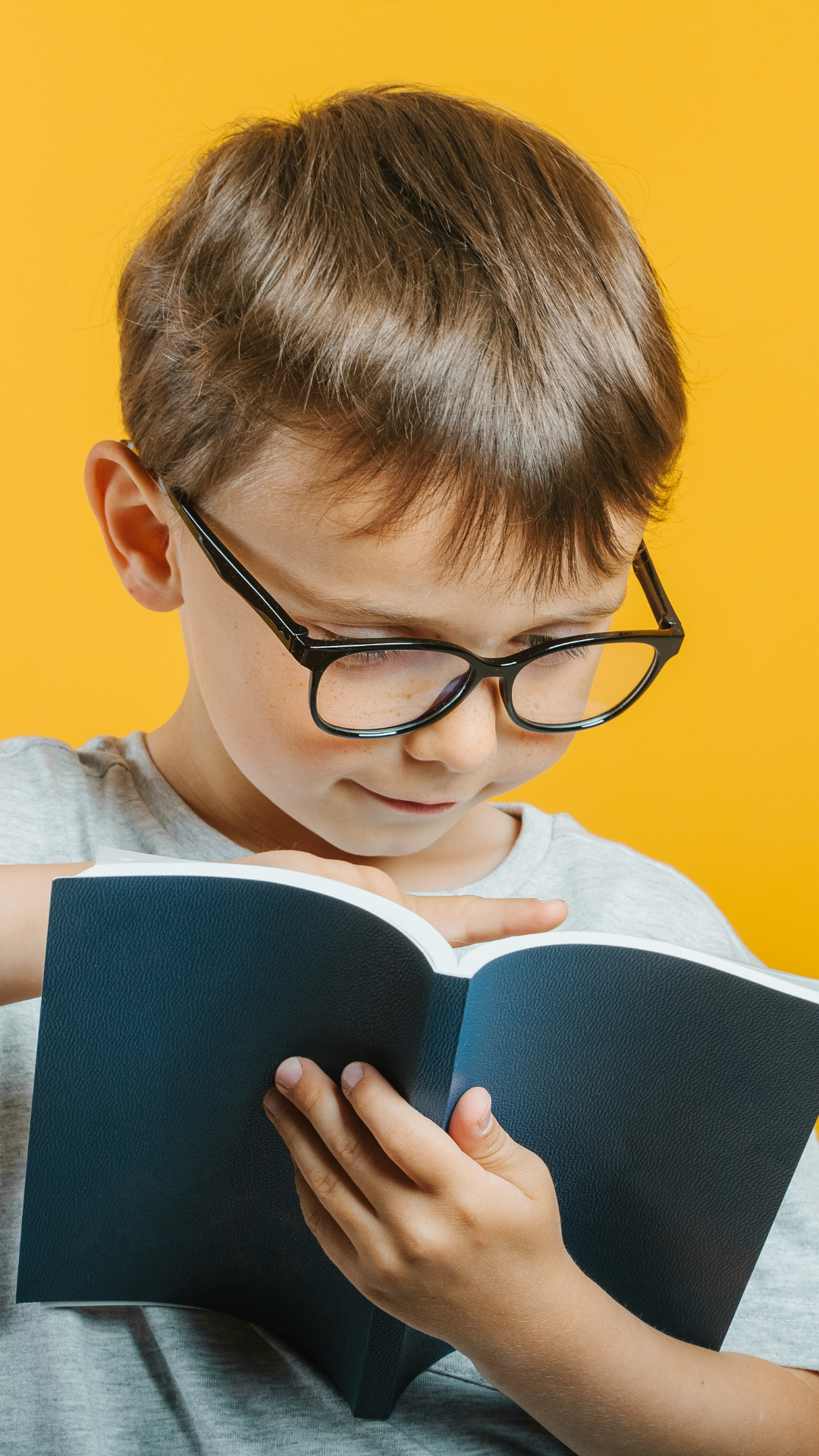 Young boy wearing glasses, reading a book against a yellow background.