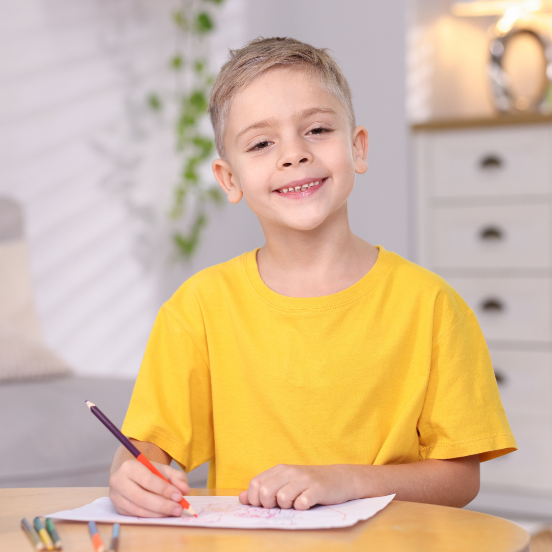 A young boy wearing a yellow t-shirt sitting at a table, smiling, and drawing with colored pencils.