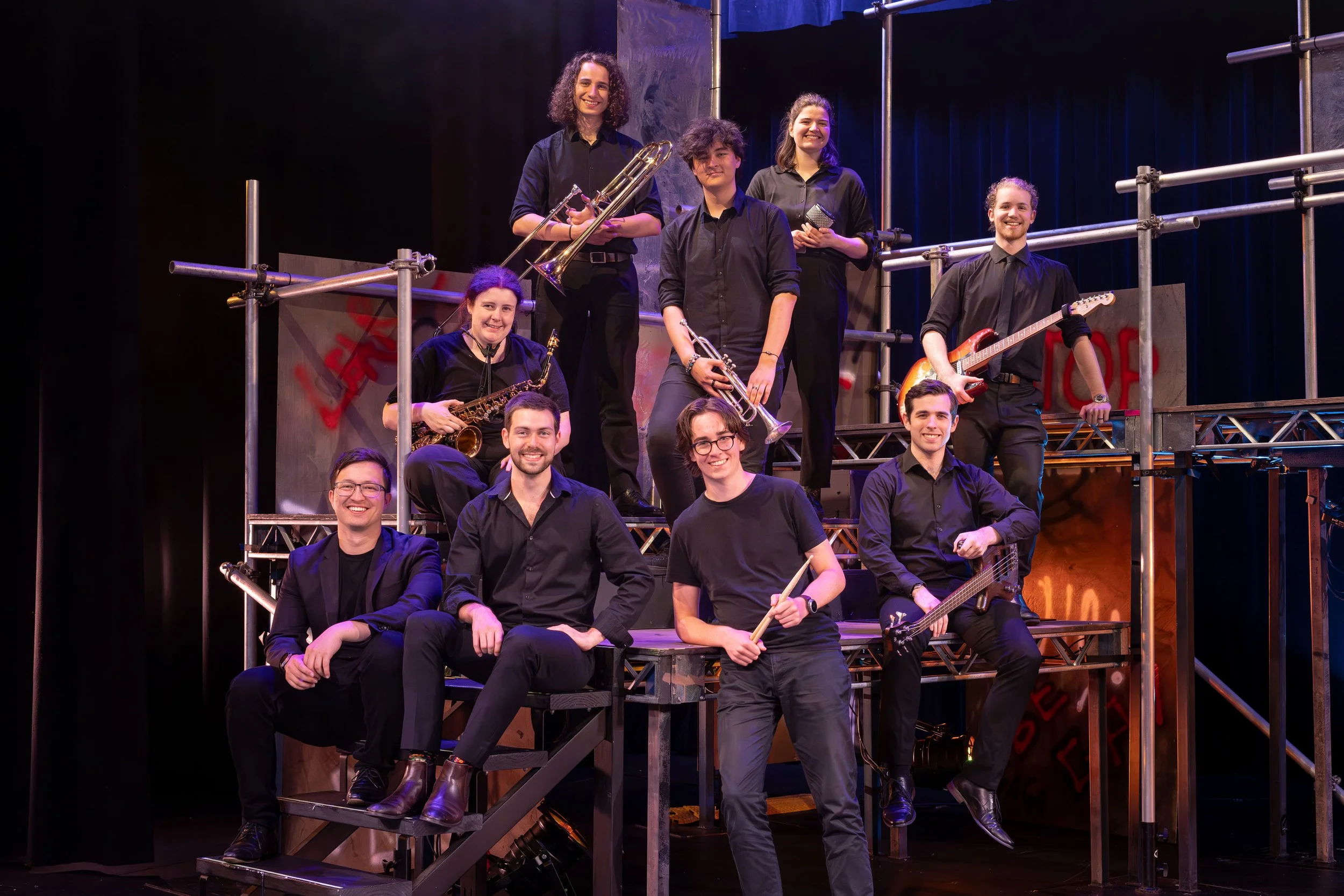 A group of ten young adults on a stage, holding musical instruments including saxophones, trumpets, electric guitar, bass, and drums, posing for a band photo with a theatrical set background.