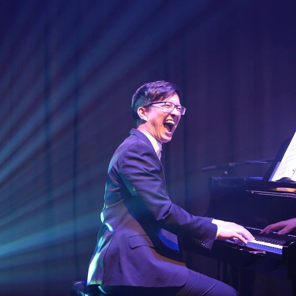A man wearing glasses and a suit playing a grand piano laughing while playing on a stage with purple lighting.