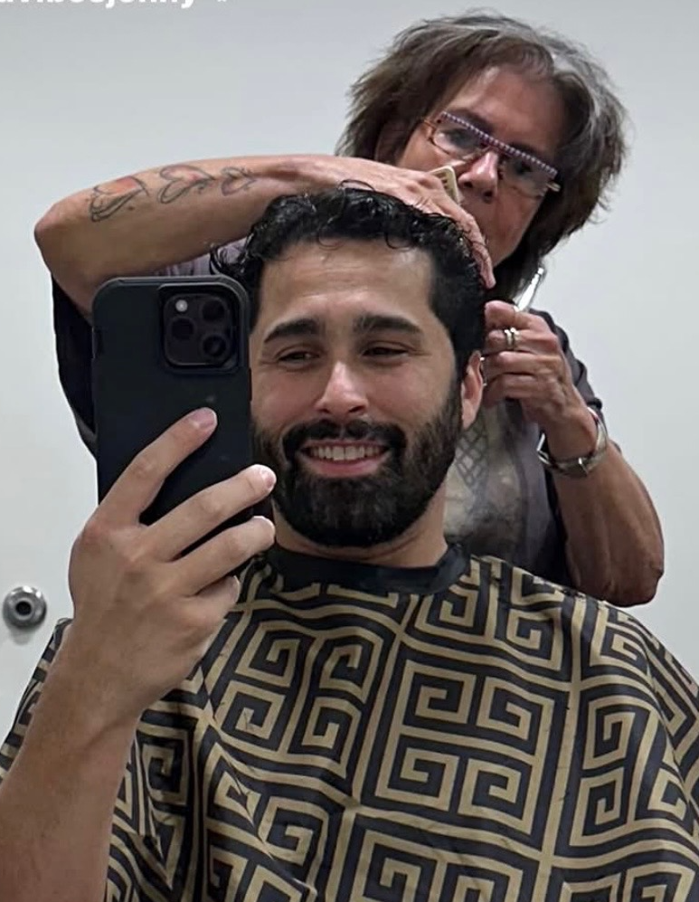 Man with dark hair and beard taking a selfie in a barber chair, smiling, with a hairstylist behind him service styling his hair in a salon.