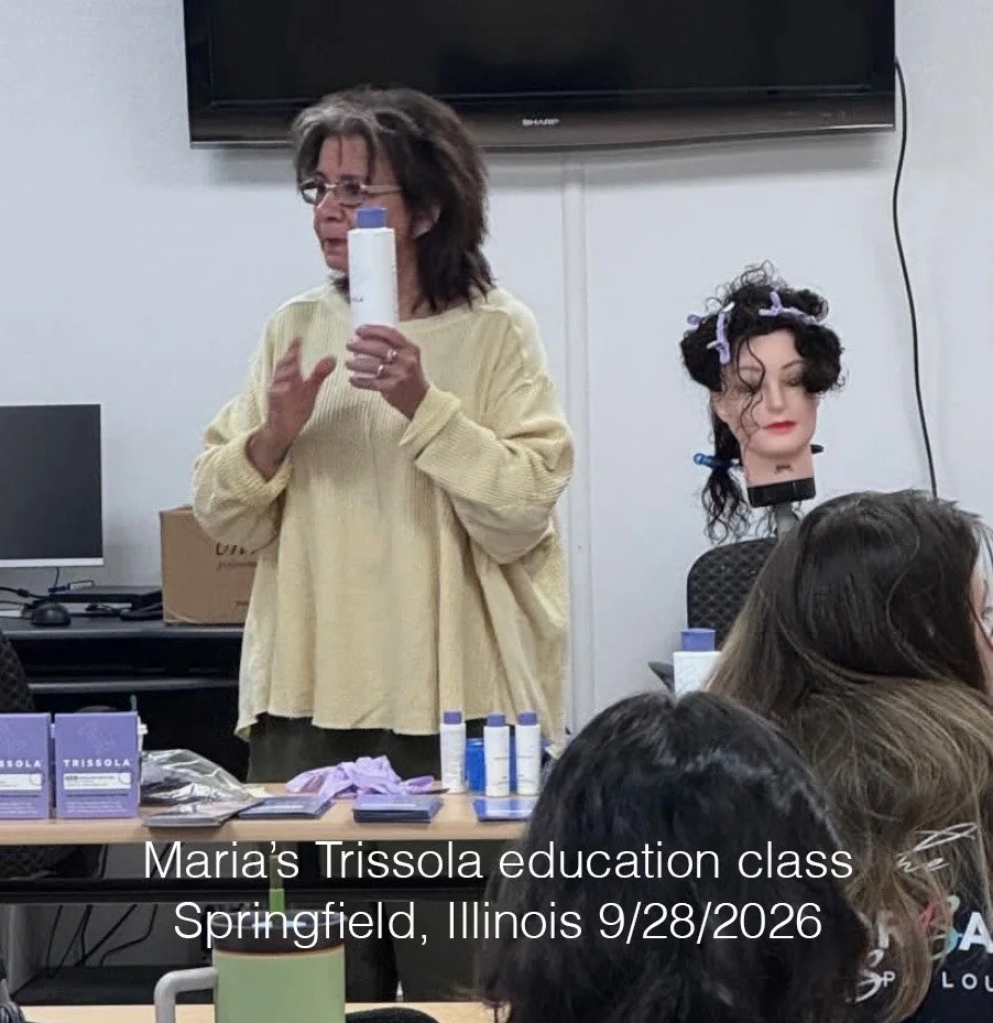 A woman in a yellow sweater demonstrating hair product usage in a classroom with a mannequin head with styled hair and students sitting in front, in Springfield, Illinois, on September 28, 2026.