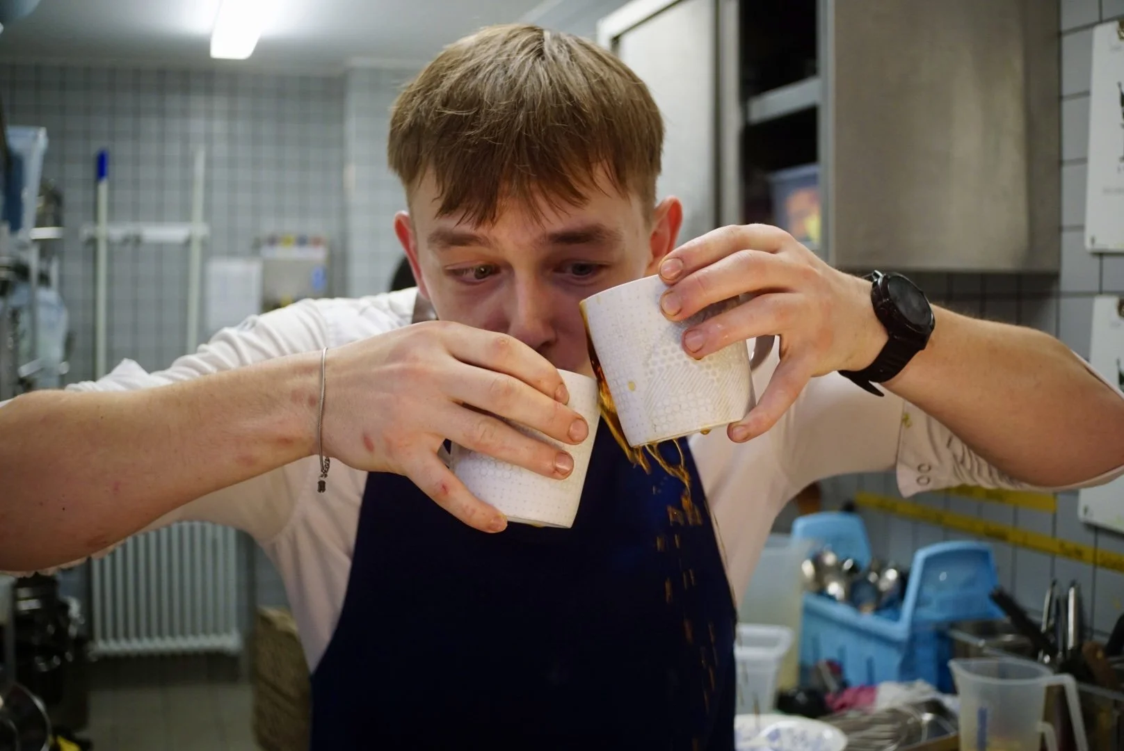 A man with messy hair and a black watch on his left wrist, wearing a white shirt and apron, is holding two coffee cups, spilling coffee while pouring from one to the other in a kitchen environment.
