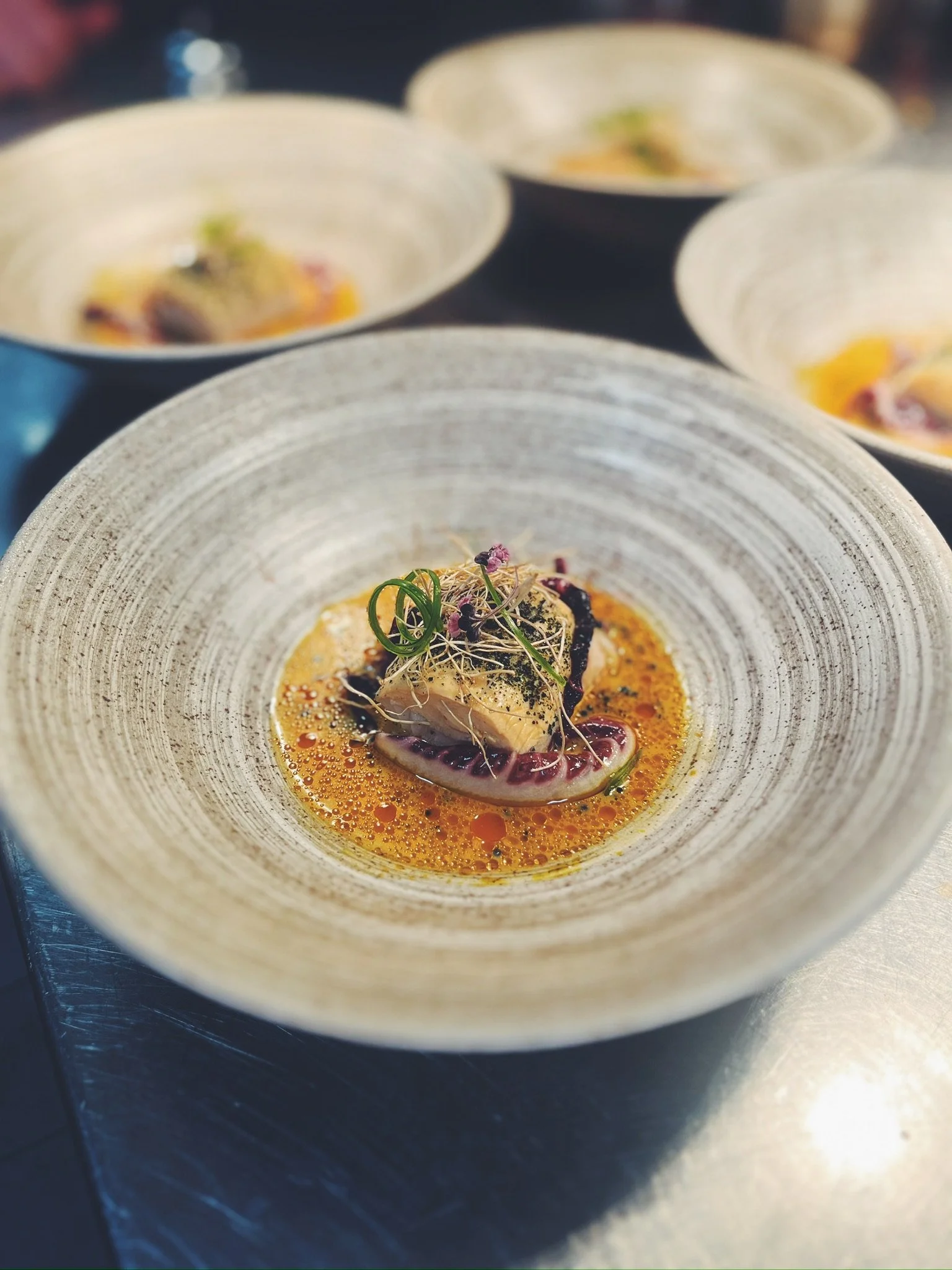 A close-up of a gourmet seafood dish with a fish fillet topped with microgreens, served in a textured bowl with a citrus sauce, butter, and oil.
