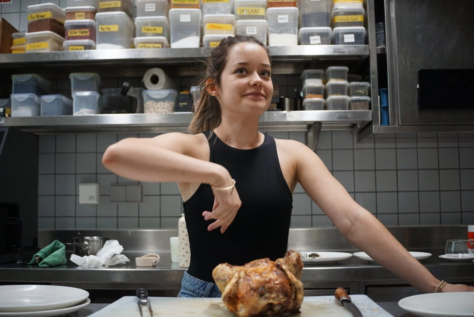 A woman in a black sleeveless top standing in a kitchen with shelves of labeled containers behind her. She is posing with a partially carved roasted chicken on a cutting board in front of her, with a carving knife nearby.