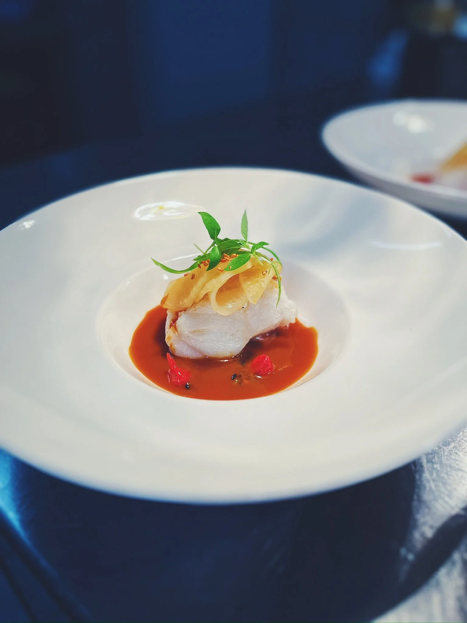 A gourmet dish featuring a piece of white fish topped with greens and thinly sliced vegetables on a bed of reddish-brown sauce, served in a white bowl.