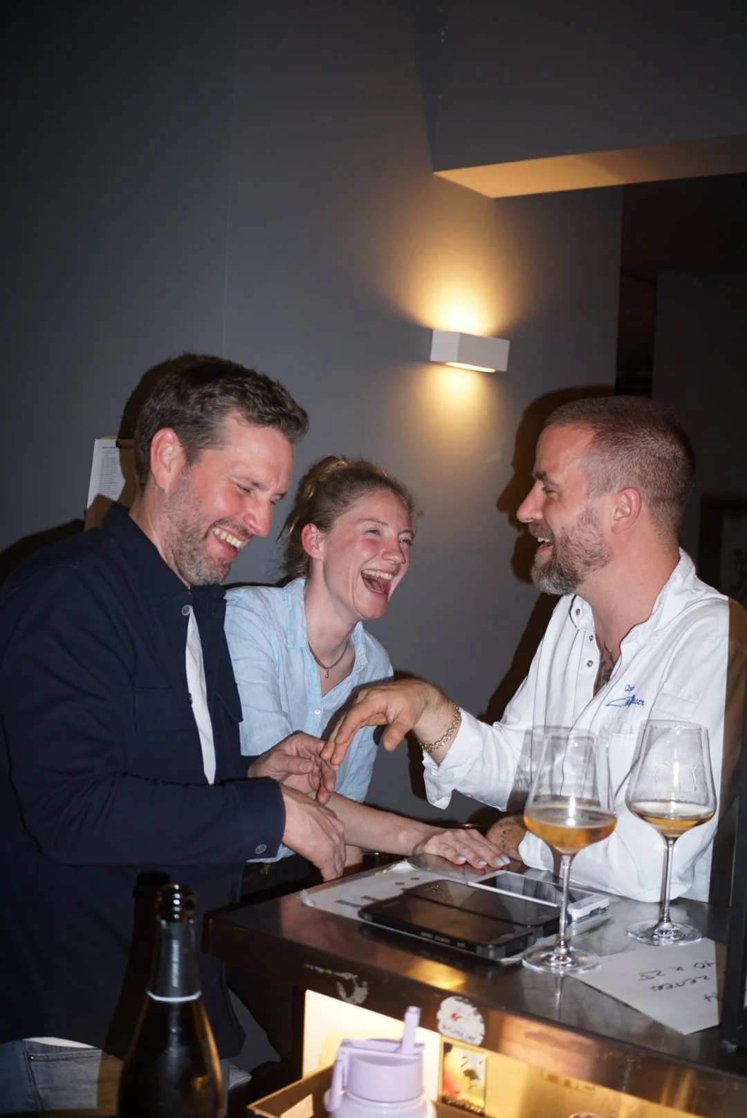 Three friends laughing and talking at a bar, with two glasses of wine or champagne on the counter.