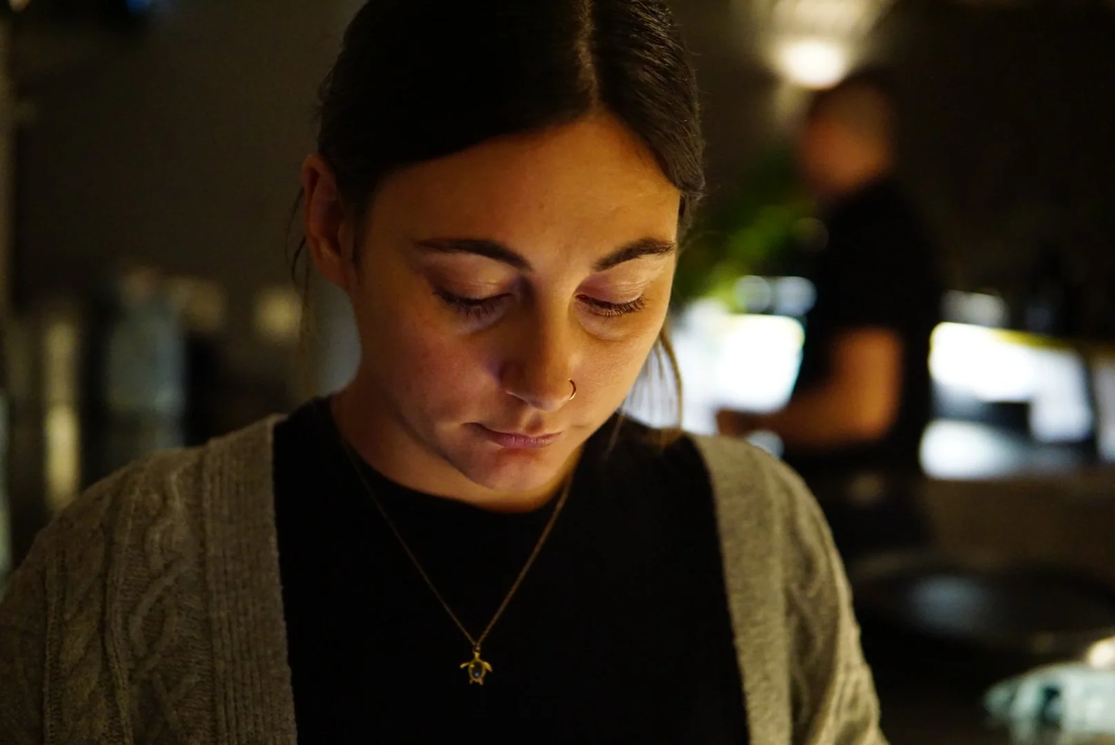 A young woman with dark hair, wearing a black top and gray cardigan, looking down with closed eyes in a dimly lit indoor setting. She wears a gold necklace with a star-shaped pendant and has a nose ring.